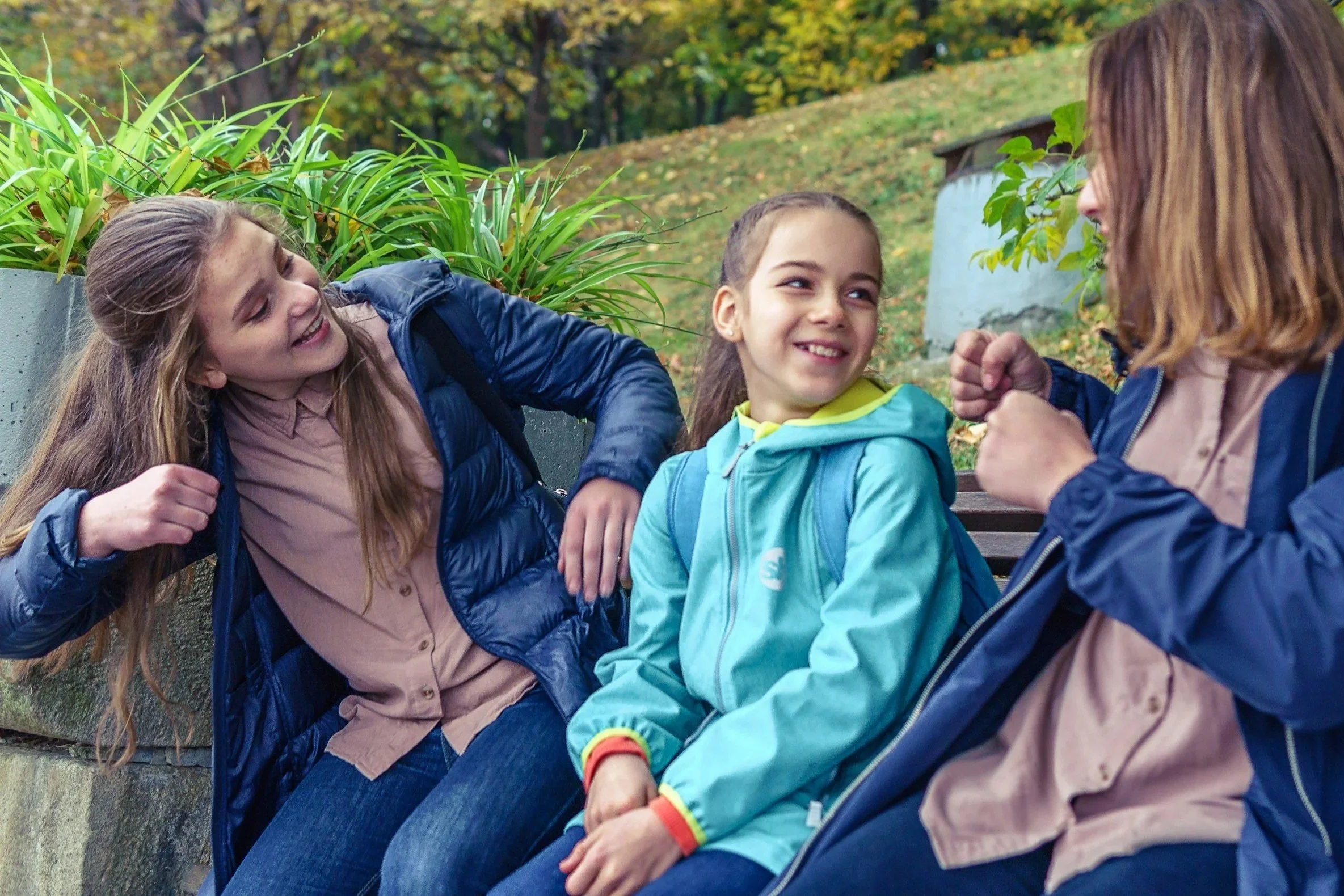 Three girls sitting on a park bench, engaged in conversation and smiling, with fall foliage in the background.