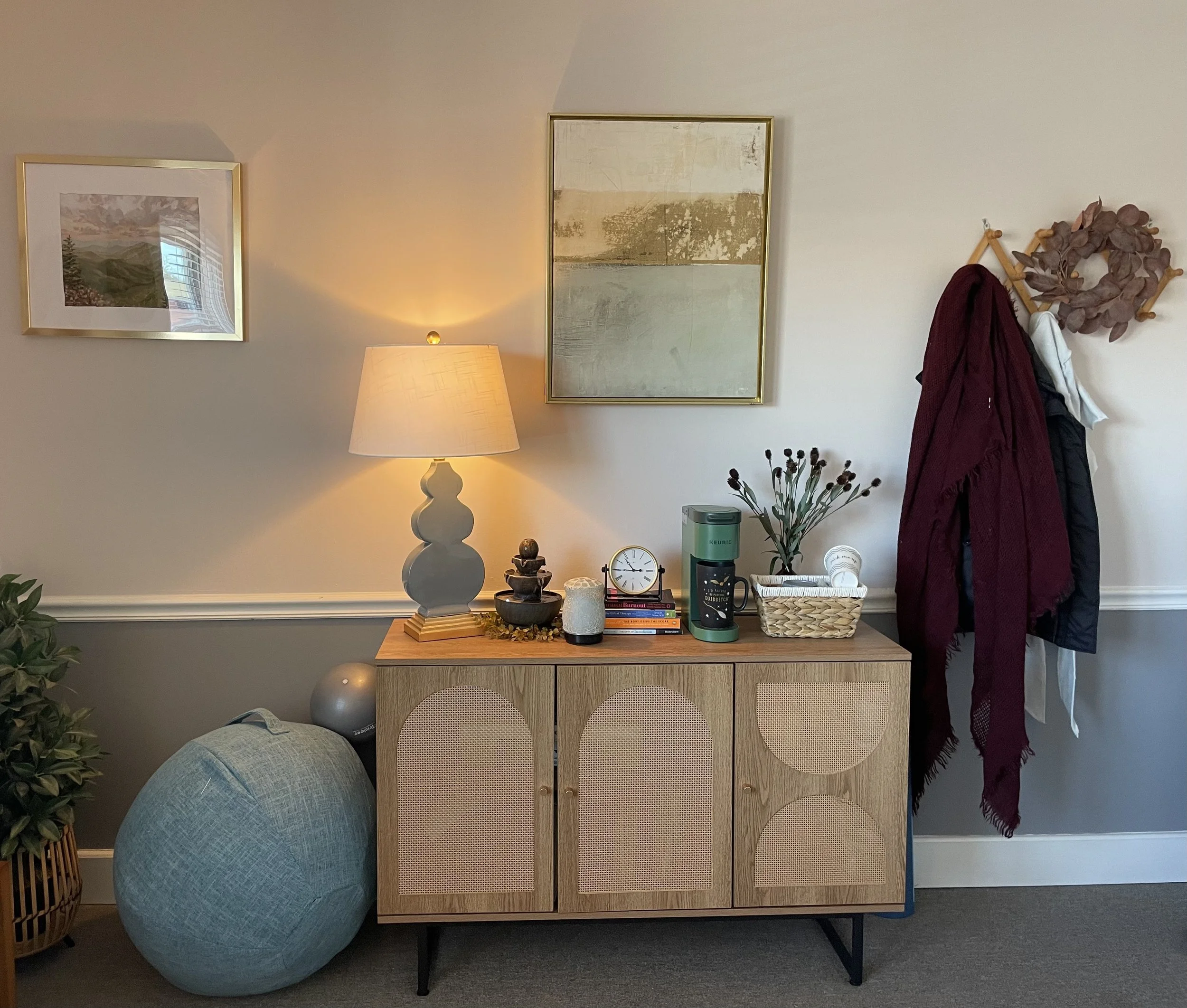 A wooden sideboard with rattan doors topped with a lit table lamp, potted plant, clock, coffee mug, and other decorative items. A dartboard with fabric tied around it and coats hanging on a wall hook are above the sideboard, with framed artwork on th