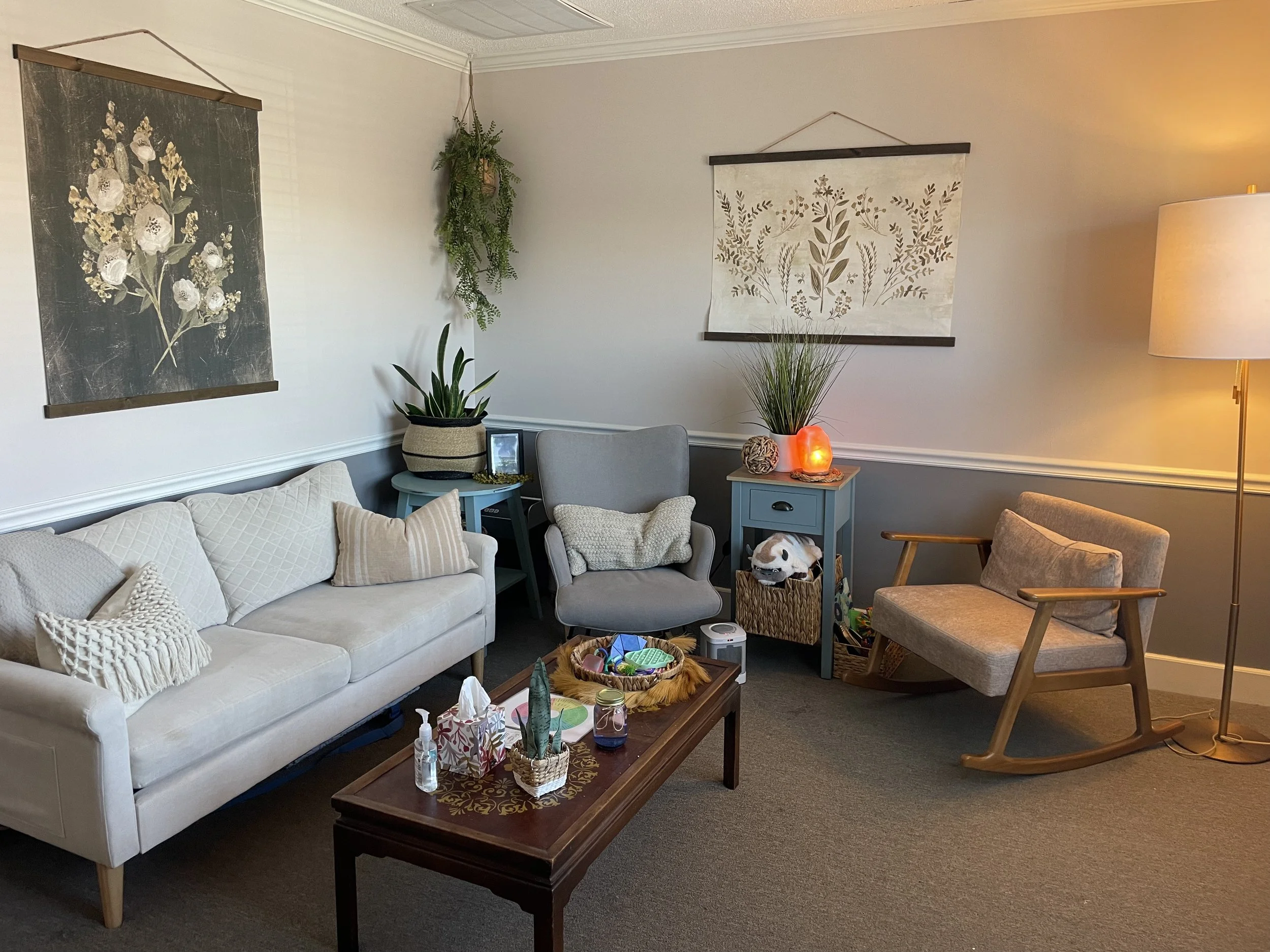 Living room with a white sofa, gray armchair, beige rocking chair, and blue side tables. Decor includes framed botanical art, hanging plant, and salt lamps. Coffee table has tissue box, decor, and hand sanitizer. Carpeted floor with a warm, cozy sett
