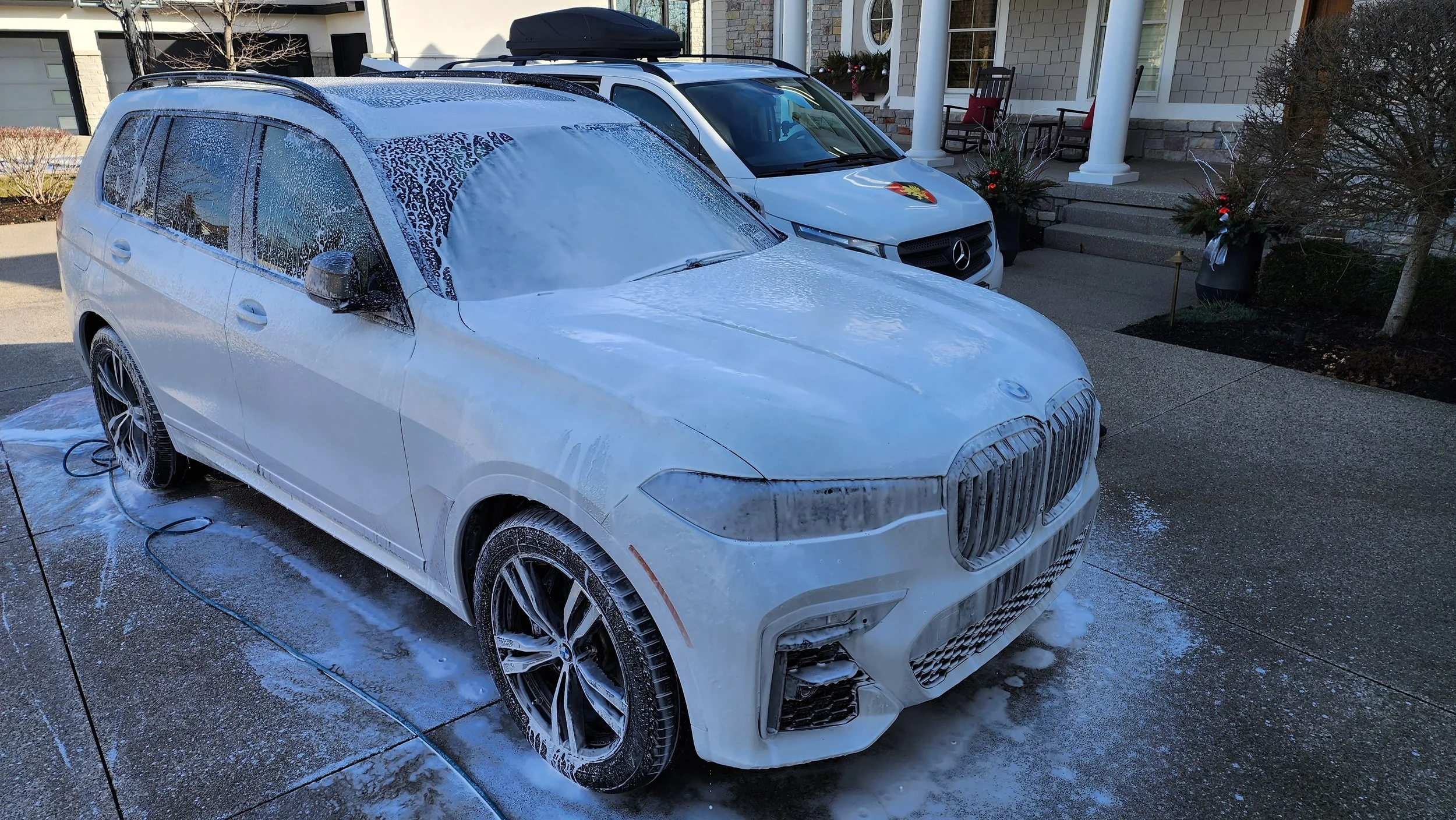 White BMW SUV being washed with soap and water on a driveway. There are two parked vehicles and a house with Christmas decorations in the background.