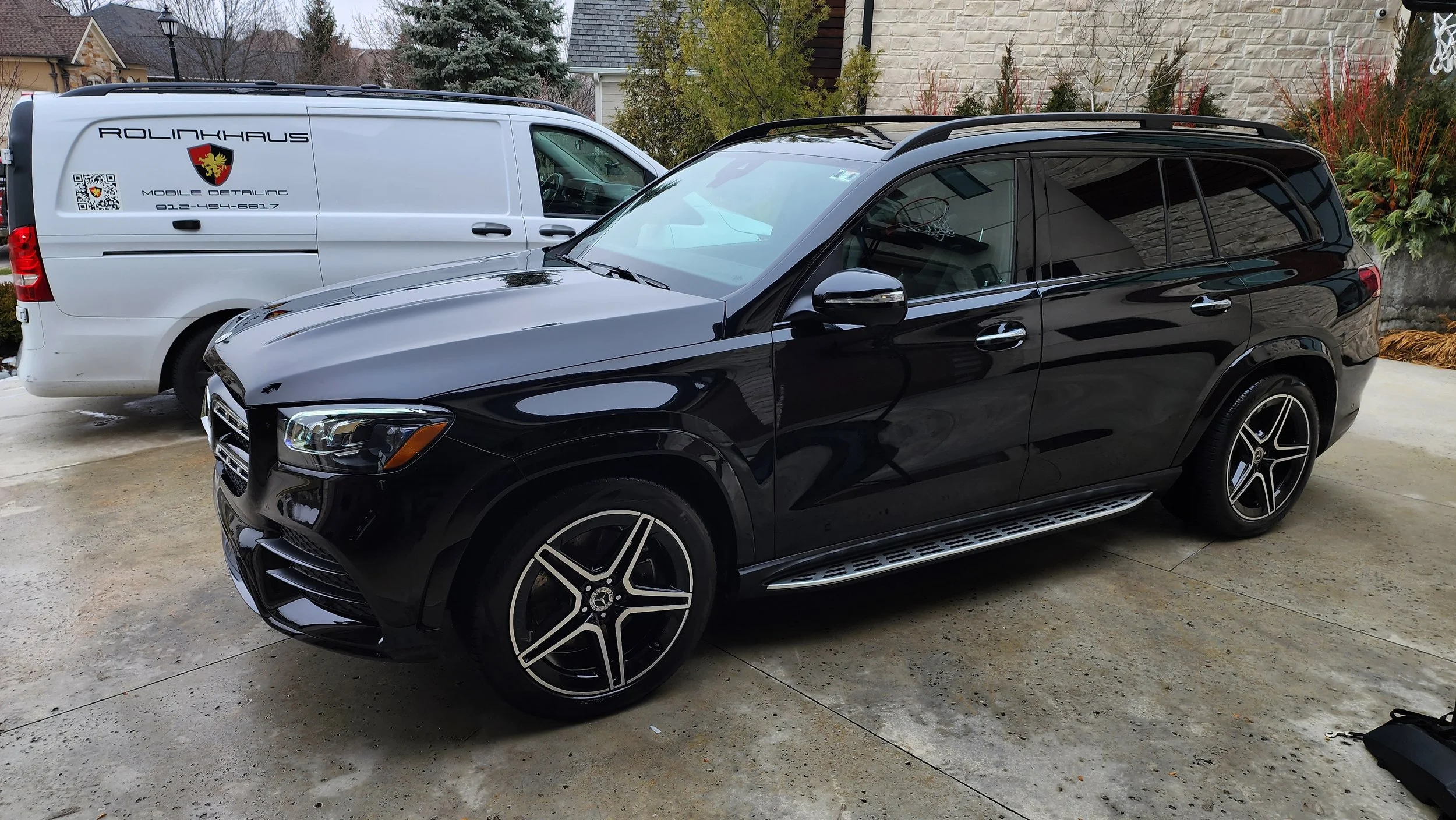 A black Mercedes-Benz SUV parked on a driveway next to a white van labeled 'Rolinkhaus Mobile Detailing' with a phone number, in a residential area with trees and houses in the background.