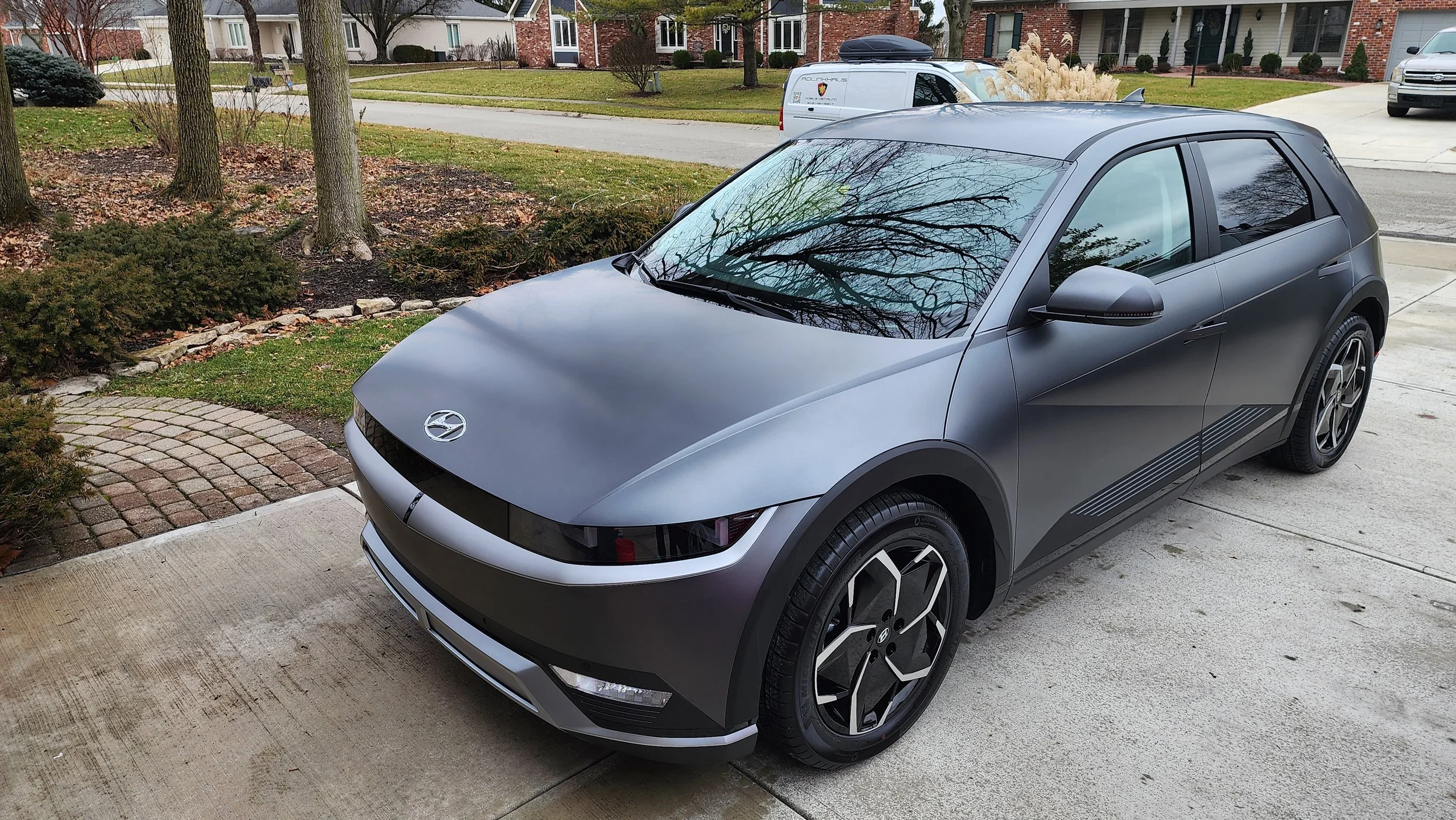 A sleek, matte gray Hyundai electric hatchback parked in a suburban driveway, reflecting leafless trees on its windshield, with a landscaped yard and a street lined with trees and houses in the background.