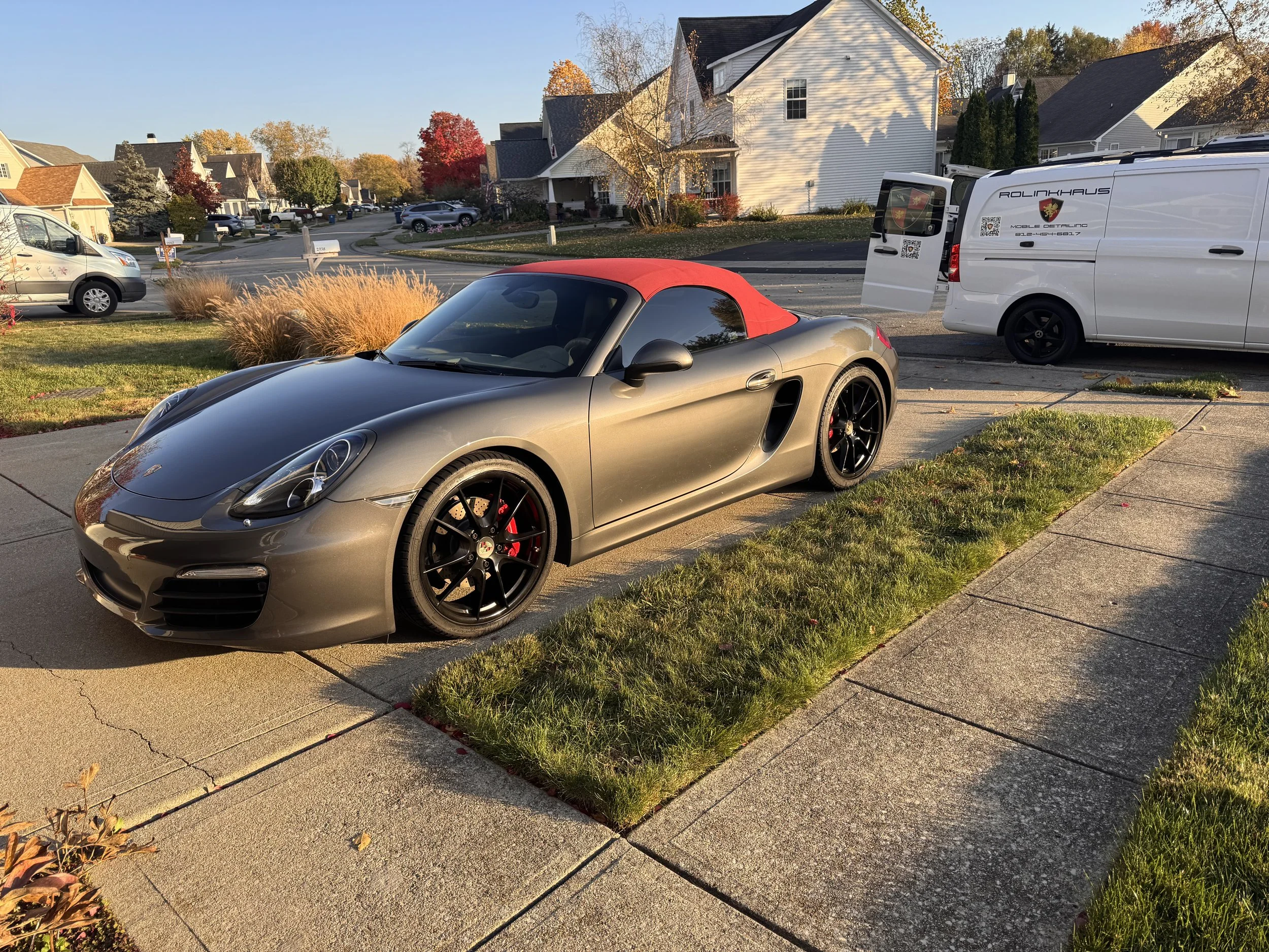 A silver Porsche convertible sports car with a red soft top parked on a residential driveway during sunset, with a white work van parked nearby and a neighborhood with houses and trees in the background.