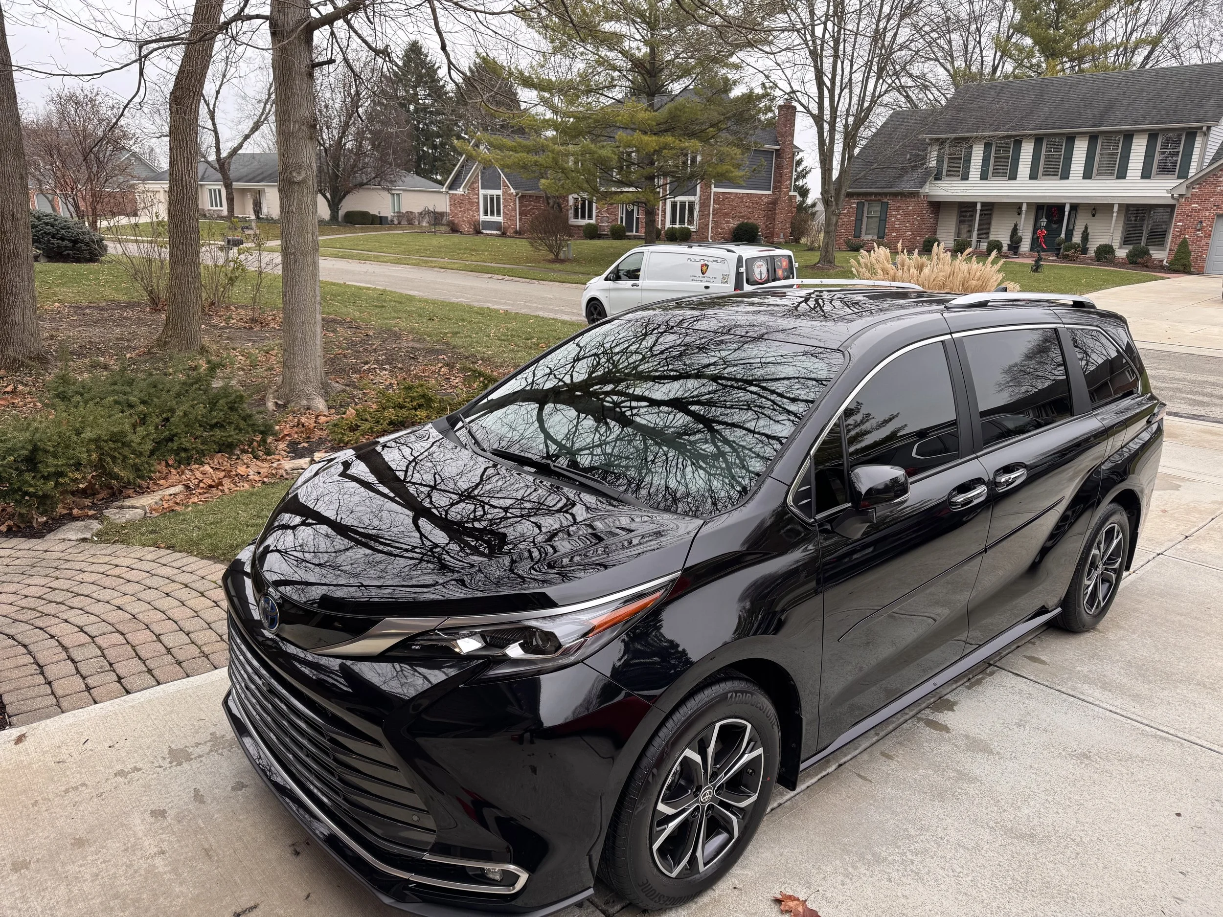Black minivan parked in driveway with trees, other houses, and a white van in the background