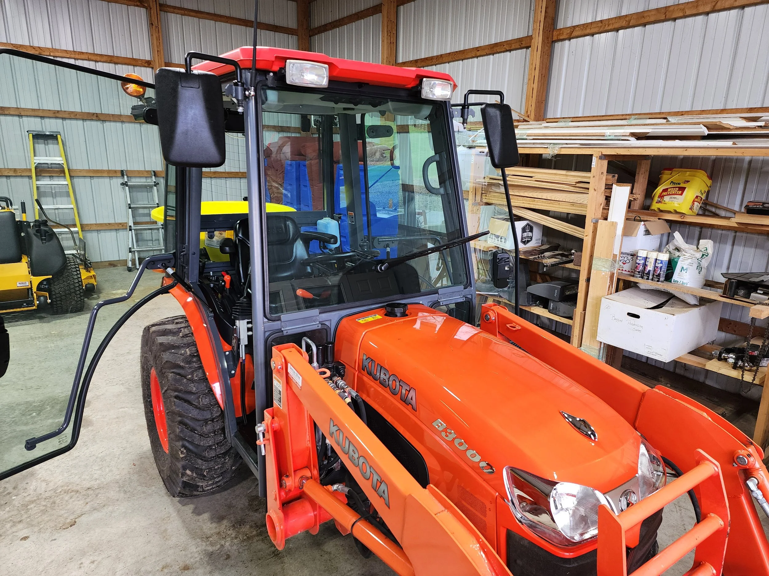 Orange Kubota tractor inside a storage shed with wooden shelves holding tools and supplies.