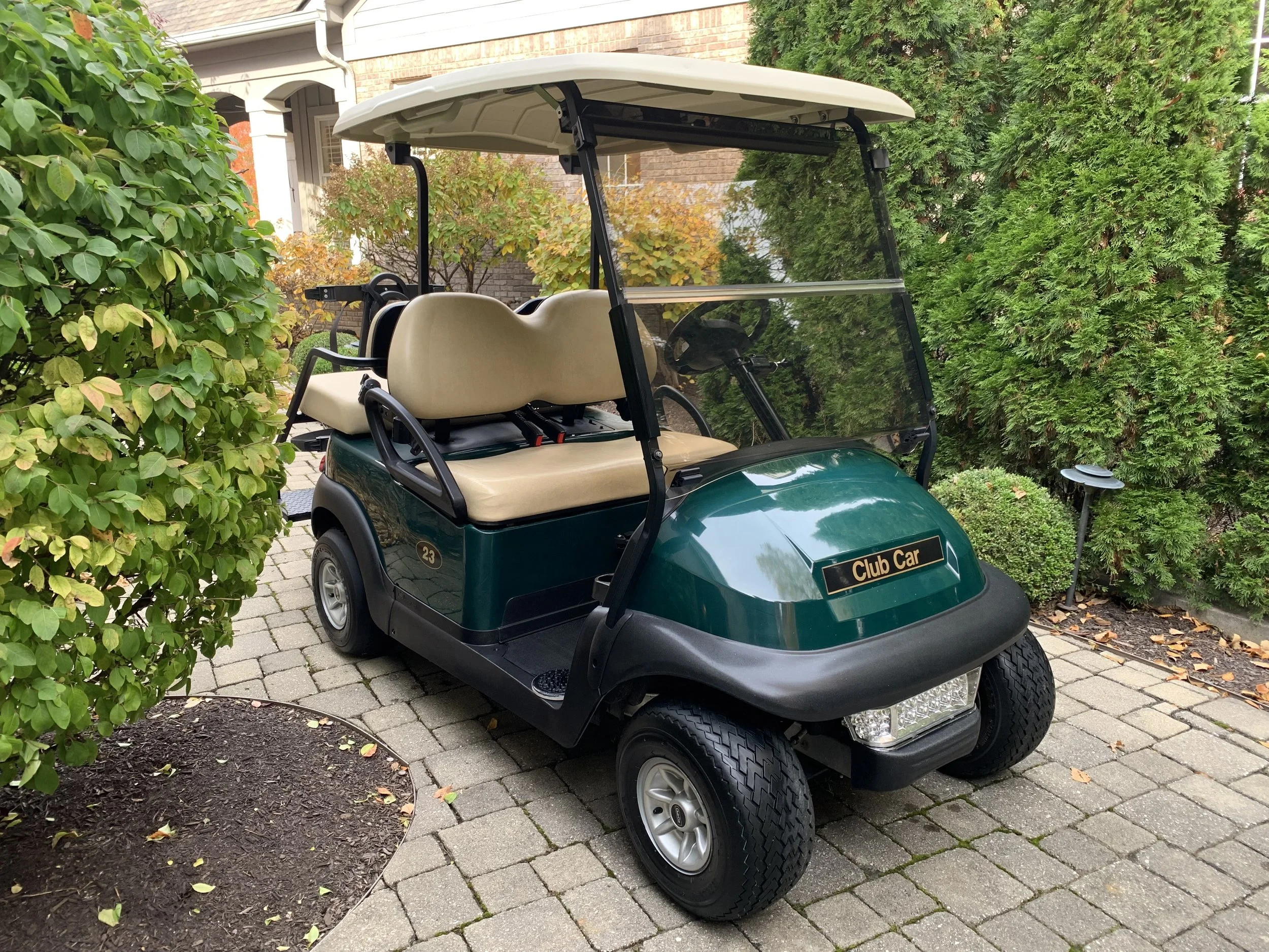 Green golf cart with a beige roof and seats, labeled 'Club Car', parked on a cobblestone pathway surrounded by bushes and trees.