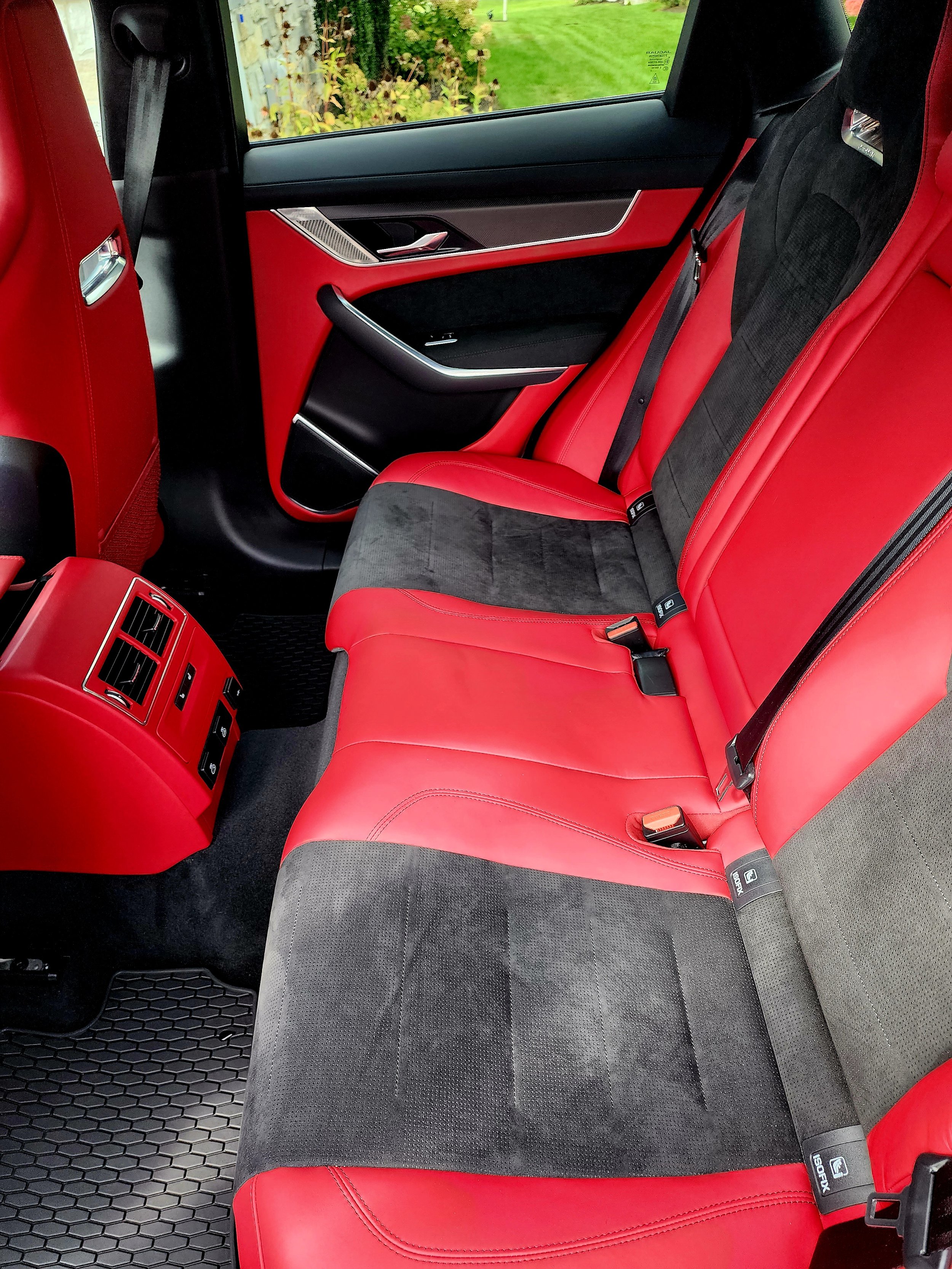 The backseat of a vehicle with red leather and black fabric upholstery, black seat belts, and a black and red door panel, with a grassy outdoor scene visible through the window.