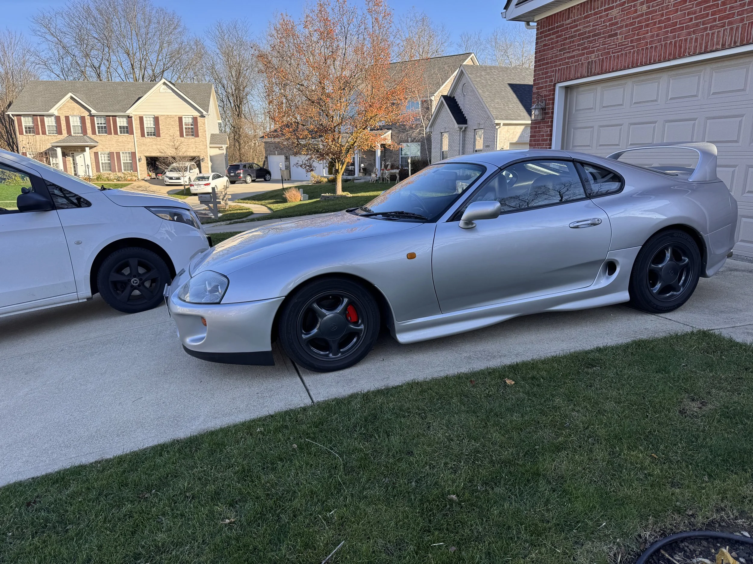 A silver sports car parked in driveway next to a white minivan, with suburban houses in the background and a tree with orange leaves.