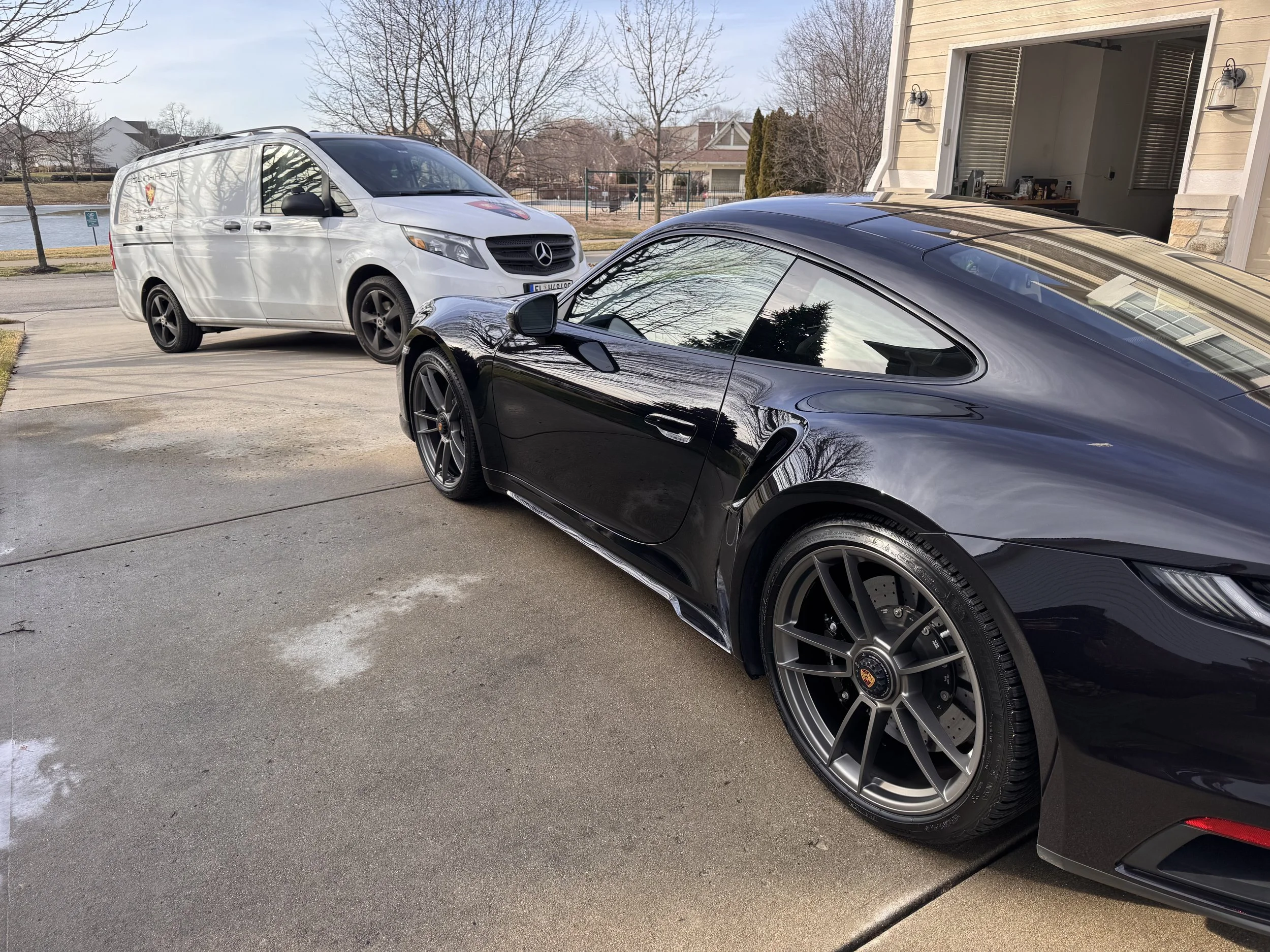A black Porsche sports car and a white Mercedes-Benz van parked in a driveway outside a house, with a lake and trees in the background.