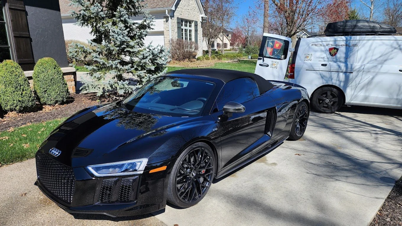 A black Audi R8 convertible parked in a driveway next to a white work van, with a house, trees, and shrubs in the background.