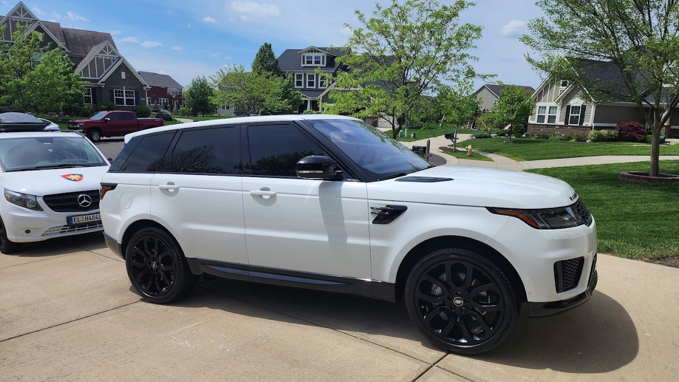 White Range Rover SUV parked on driveway with black wheels in front of a white Mercedes-Benz car in a suburban neighborhood with green lawns and trees.
