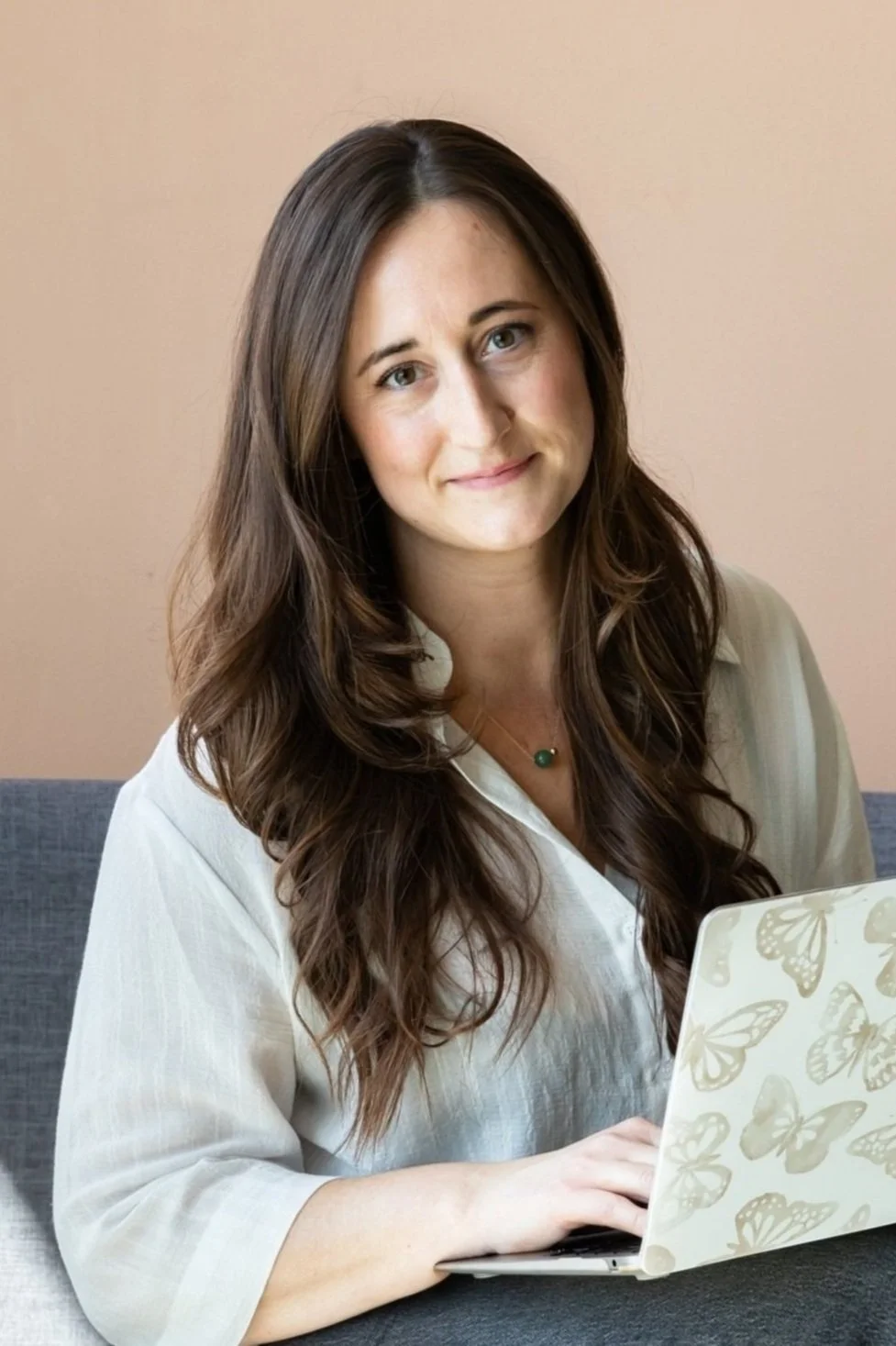 A woman with long brown hair sitting at a table using a laptop with a butterfly pattern, against a beige wall background.