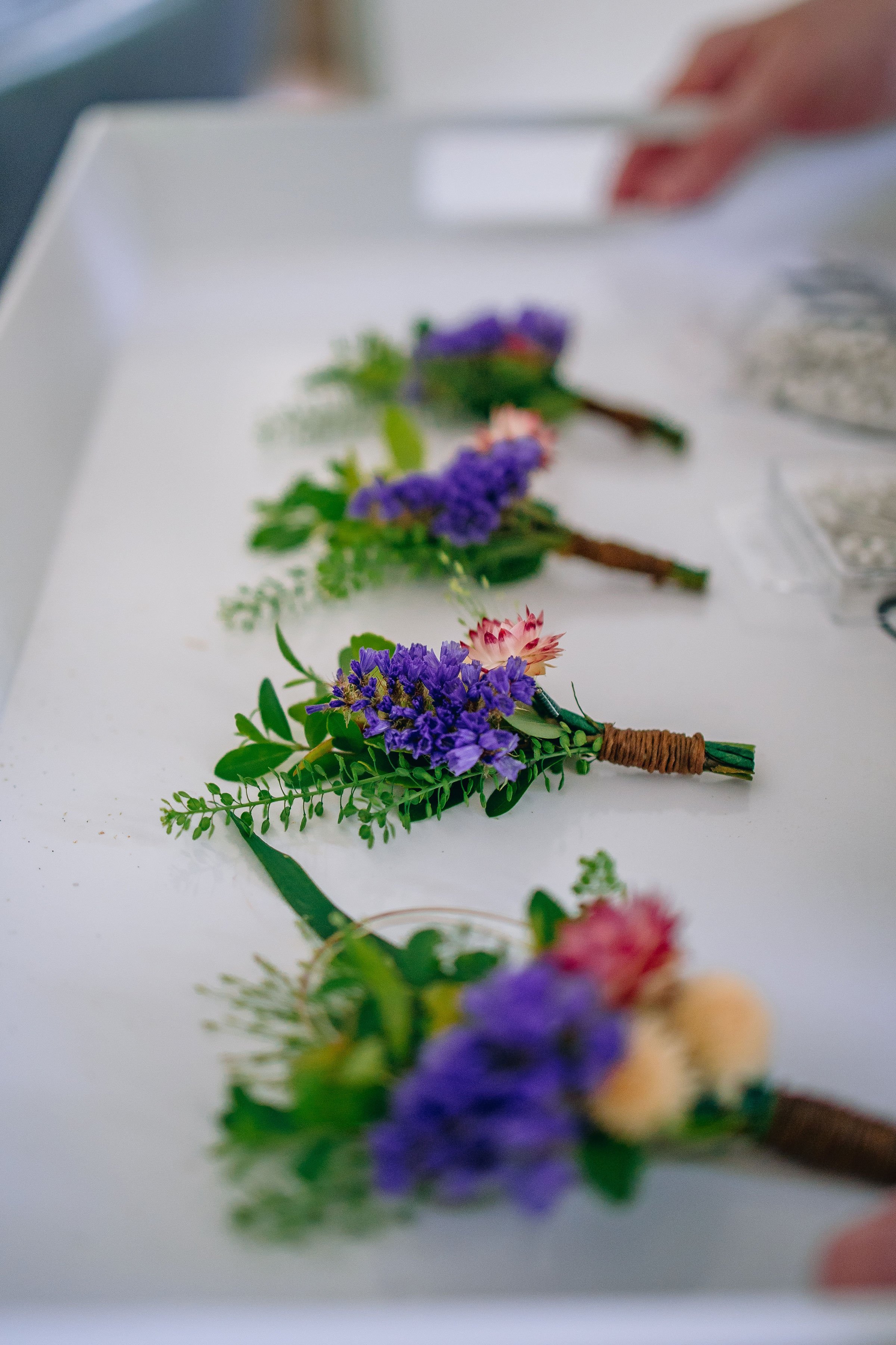 Boutonnieres for groom and groomsmen lined up on a white tray. Flowers are purple, white, and pink. Flowers by Plano, Texas florist Terra Cotta Blooms. Photo by Radiant Film &amp; Photo.