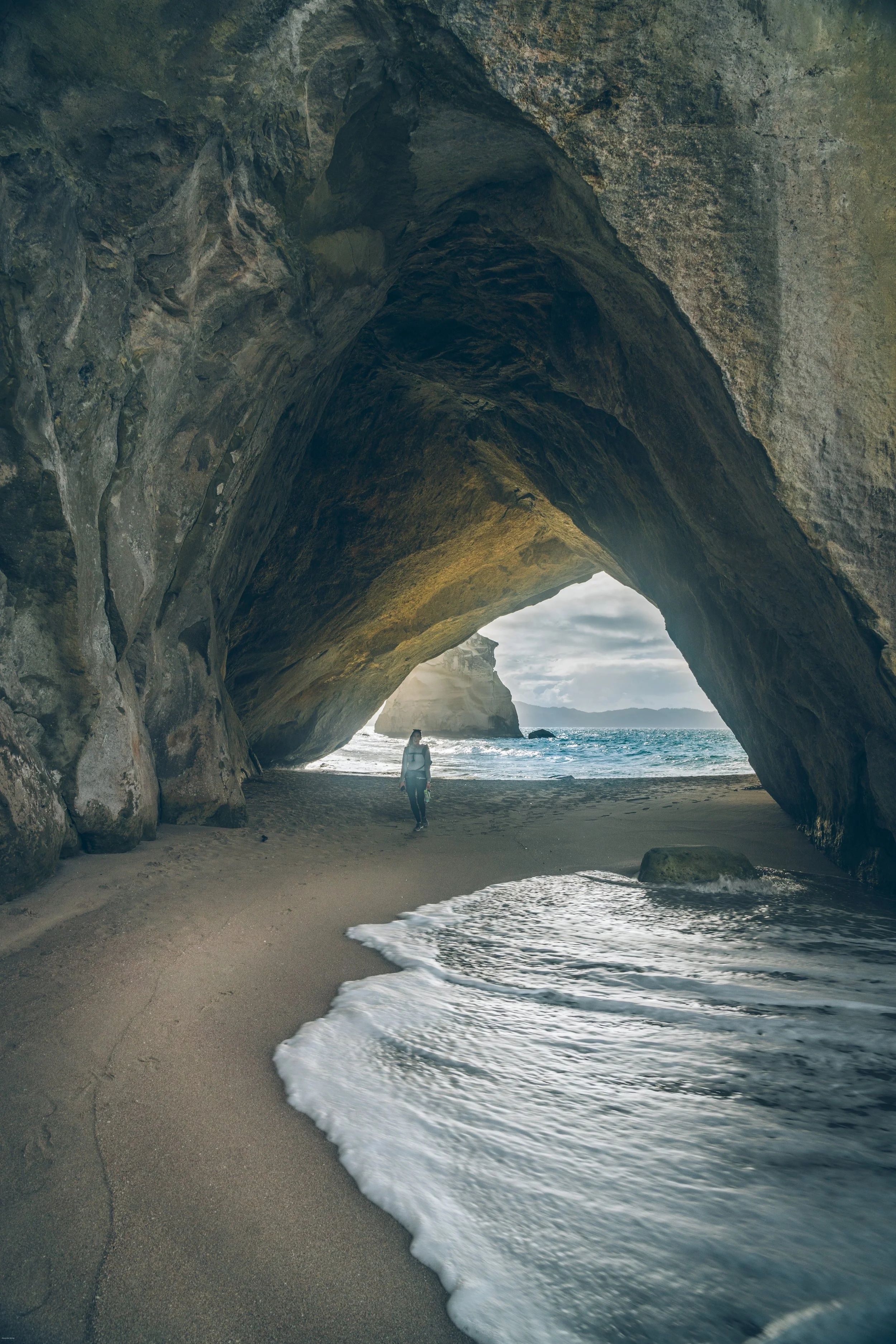 Person walking through the iconic natural rock arch at Cathedral Cove on the Coromandel Peninsula, North Island, New Zealand, as ocean waves wash across the beach.
