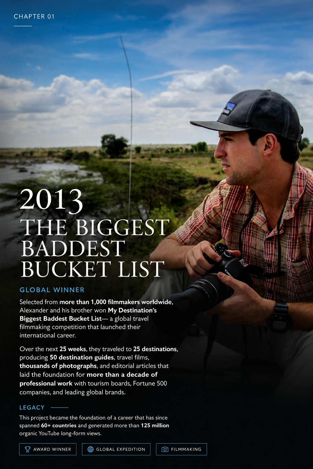 A man in a plaid shirt and baseball cap holding a camera, sitting outdoors near a river with a grassy landscape and cloudy sky in the background.