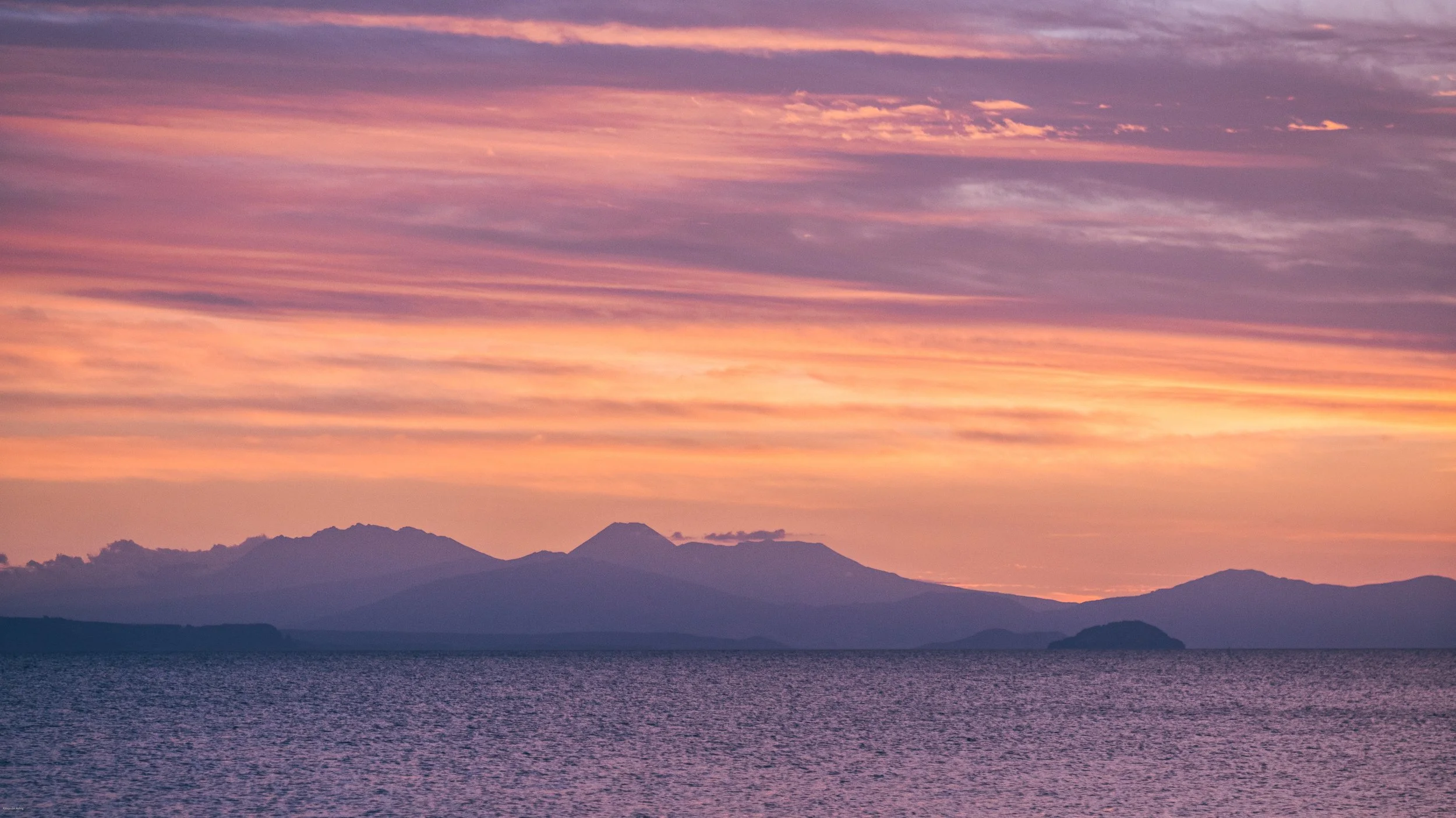 Colorful sunset over Lake Taupō with the volcanic peaks of Tongariro National Park silhouetted beneath a pink and orange sky on New Zealand’s North Island.