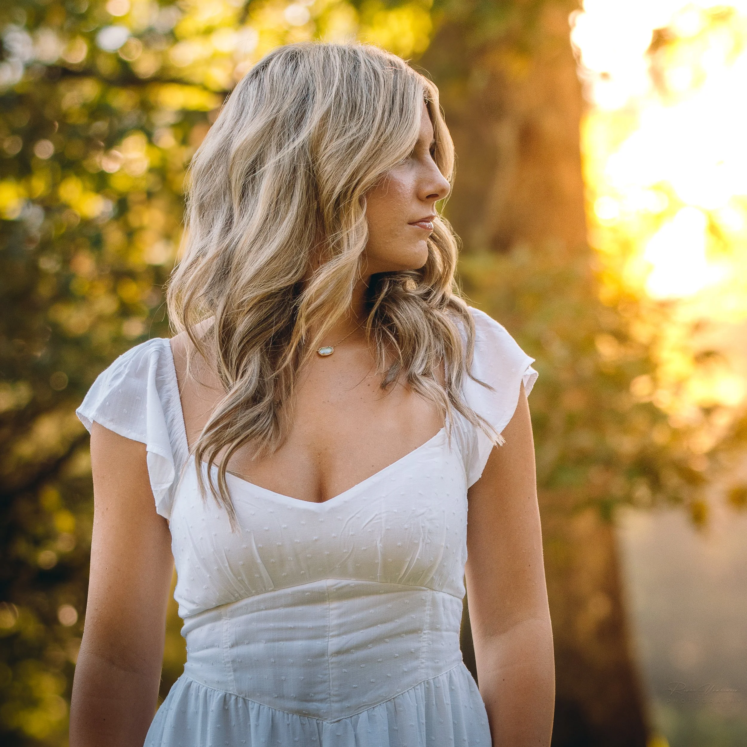 A woman with blonde hair in loose waves, wearing a white dress with ruffled sleeves, standing outdoors in a sunlit park with trees in the background.