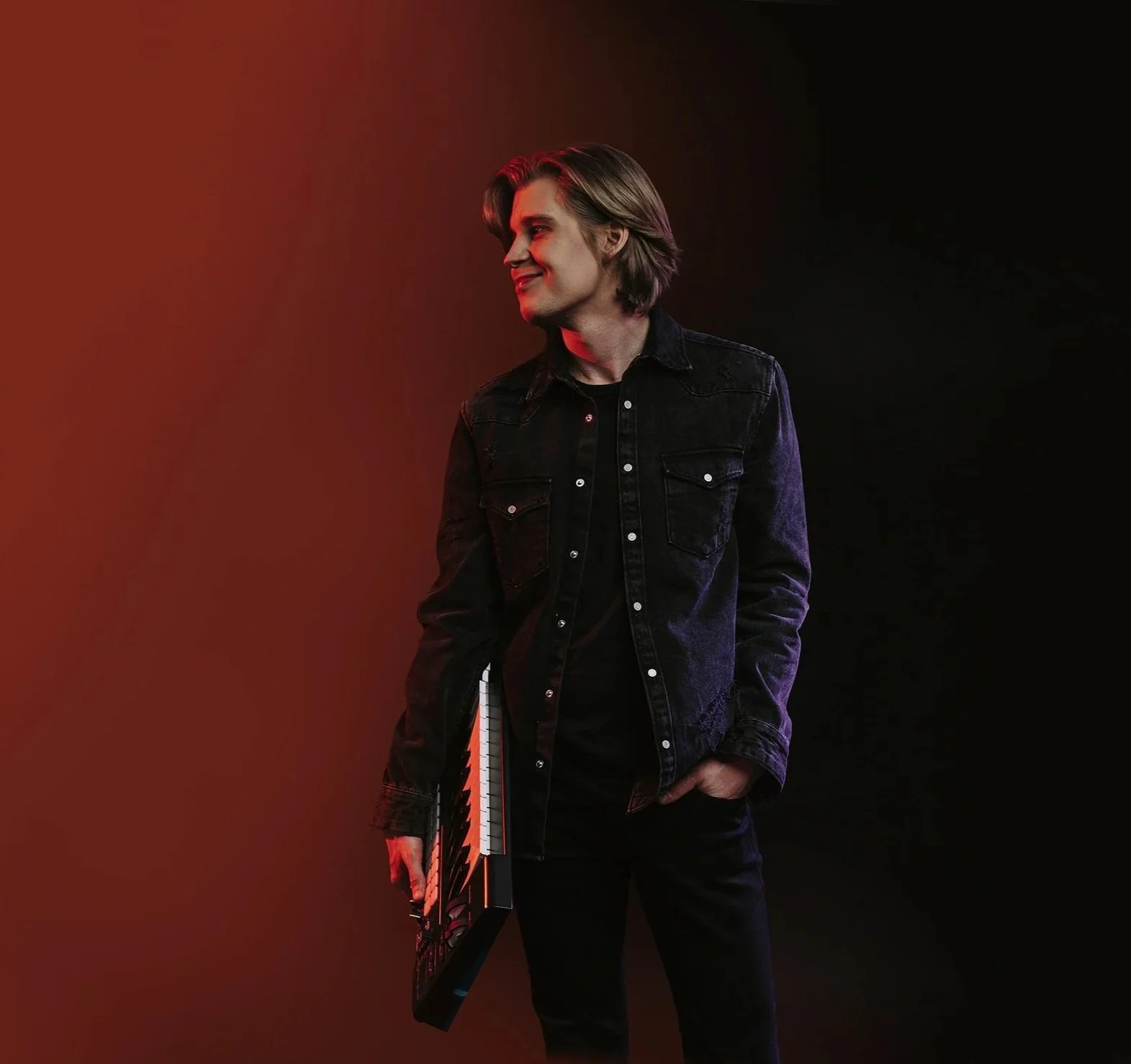 DJ and music producer holding a keyboard, photographed in studio lighting with a dark, minimalist background.