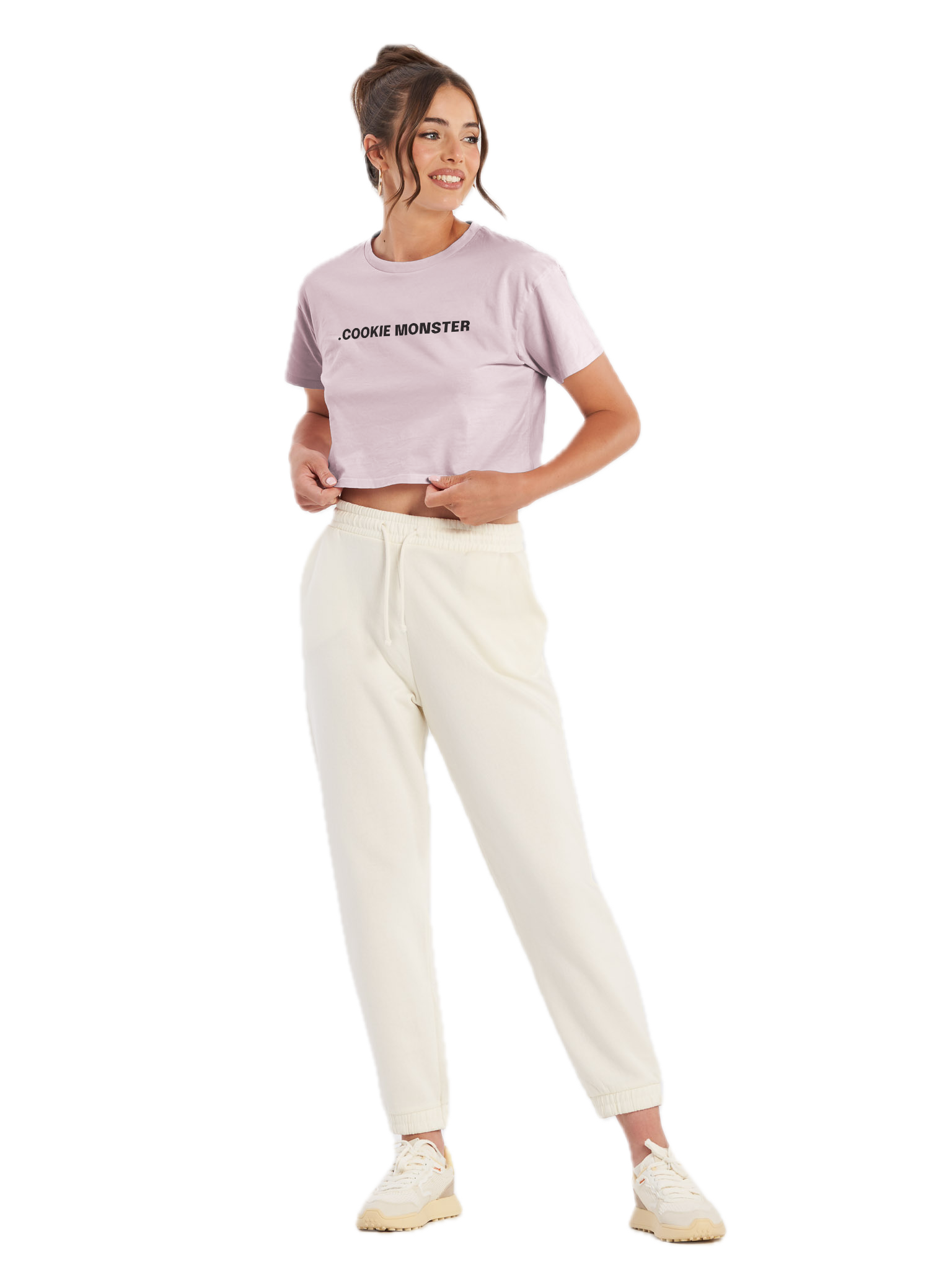 A young woman with brown hair in a ponytail, wearing a cropped pink T-shirt that says '.COOKIE MONSTER', cream-colored sweatpants, and beige sneakers, standing with one hand on her waist and the other adjusting her shirt, smiling slightly to her left.