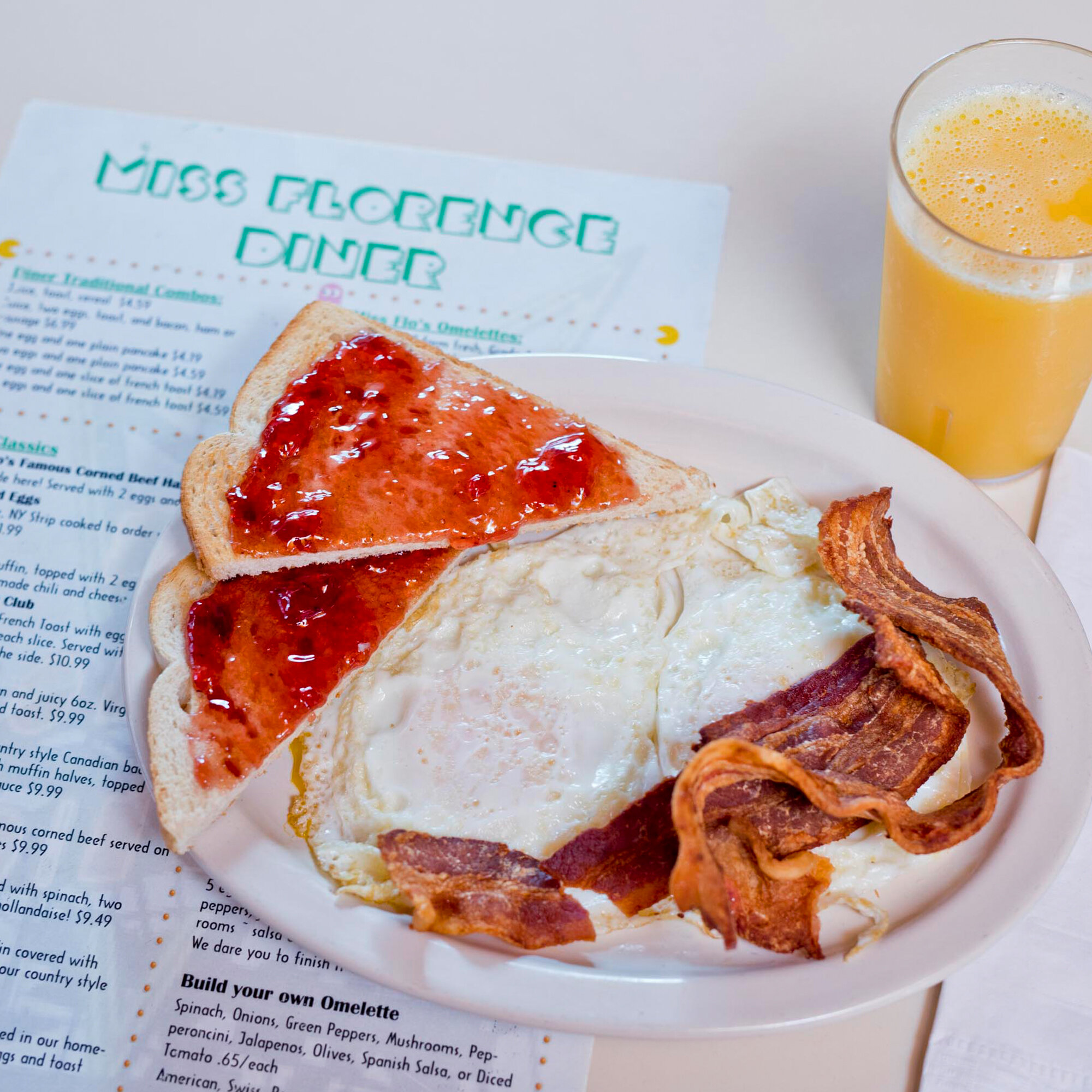  breakfast on plate including over easy eggs with a side of crispy bacon, toast with jelly and a glass of orange juice 