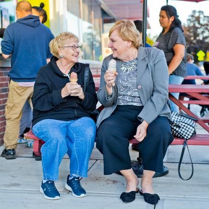  two old ladies seated side by side at a picnic table enjoying a soft serve ice cream cone after dinner on a summer night 