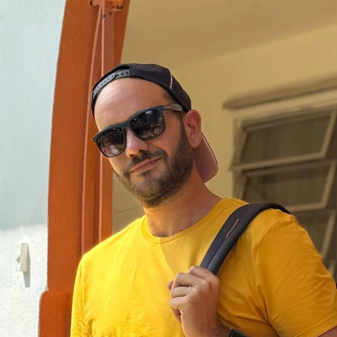 Carlos Corbeira in yellow shirt with a backpack, wearing sunglasses and a backward baseball cap, standing indoors with a slight smile.