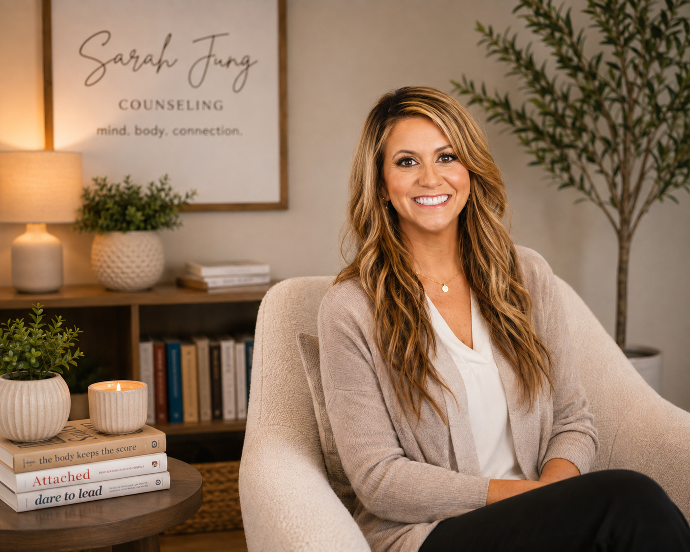 A smiling woman with long wavy hair sitting in a cozy office or living room. Behind her is a bookshelf with books, plants, and a lamp. There's a framed sign on the wall that reads 'Sarah Jung Counseling mind, body, connection.'