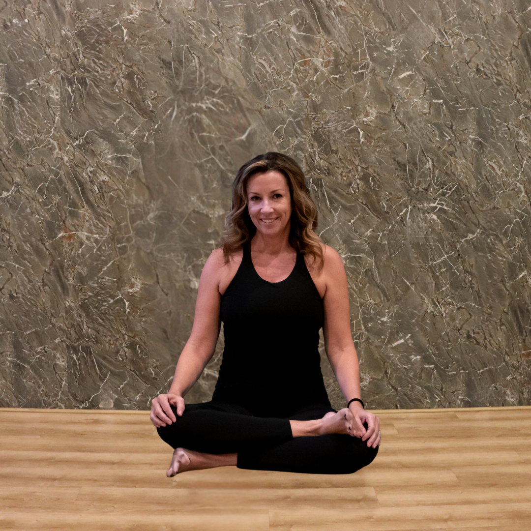 A woman practicing yoga in a seated lotus position on a wooden floor, smiling, with a textured stone wall in the background.