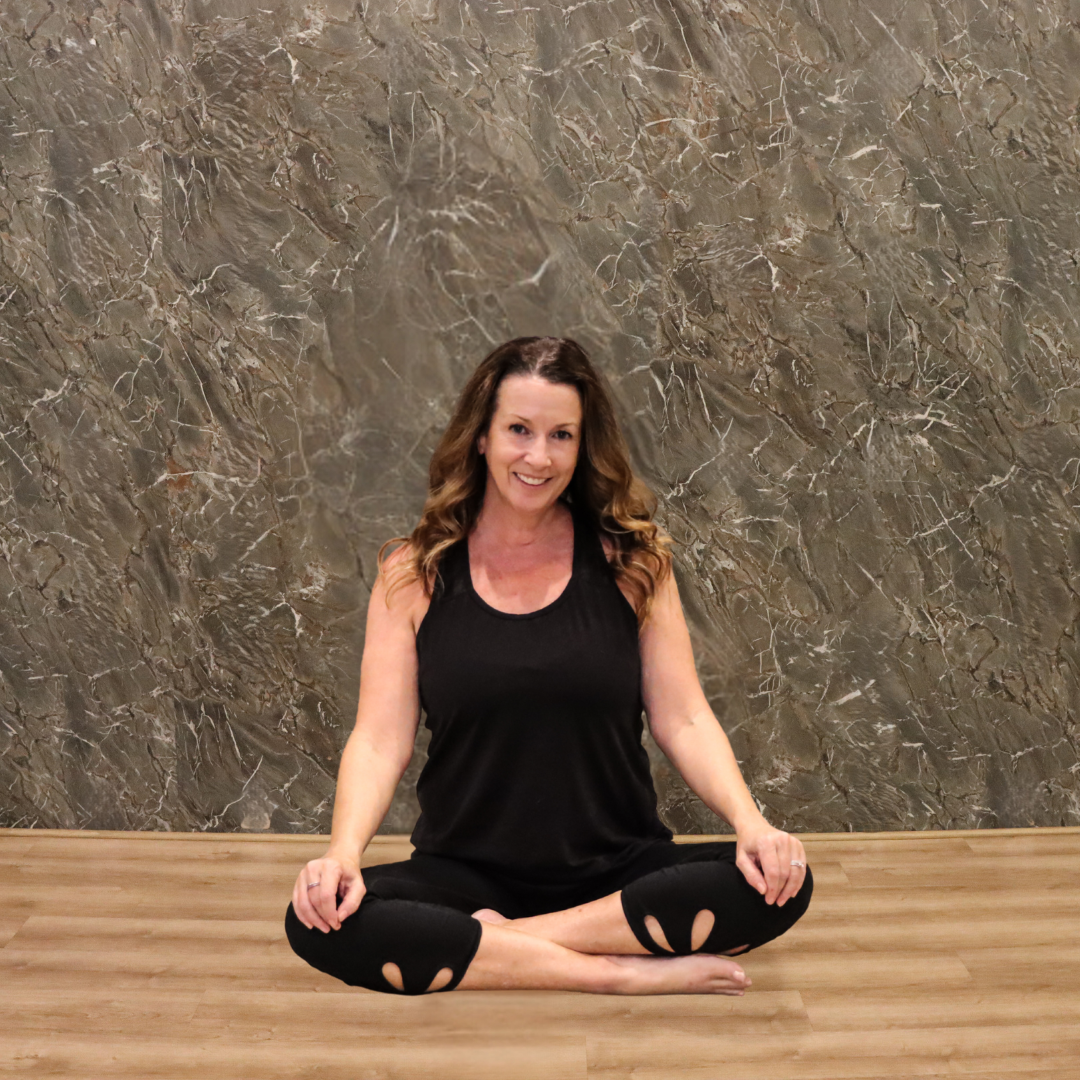 A woman practicing yoga in a seated lotus position on a wooden floor, smiling, with a textured stone wall in the background.