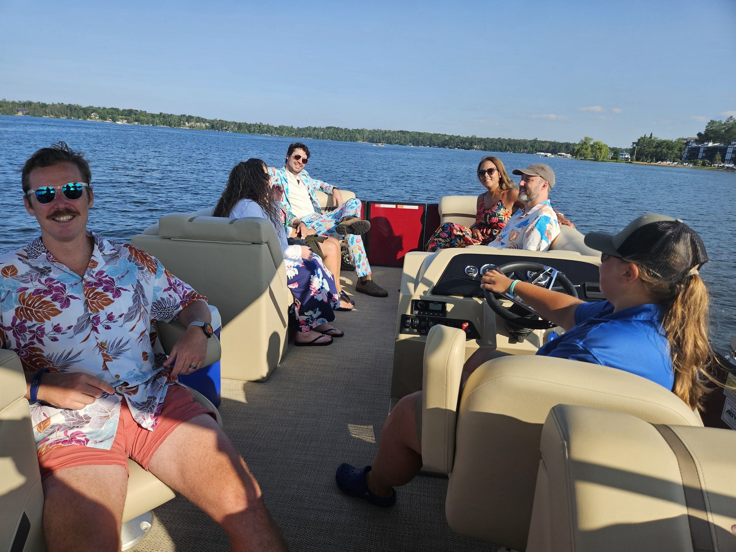 Group of people relaxing on a pontoon boat on a lake, wearing casual summer clothes and sunglasses, with a sunny sky and shoreline in the background.