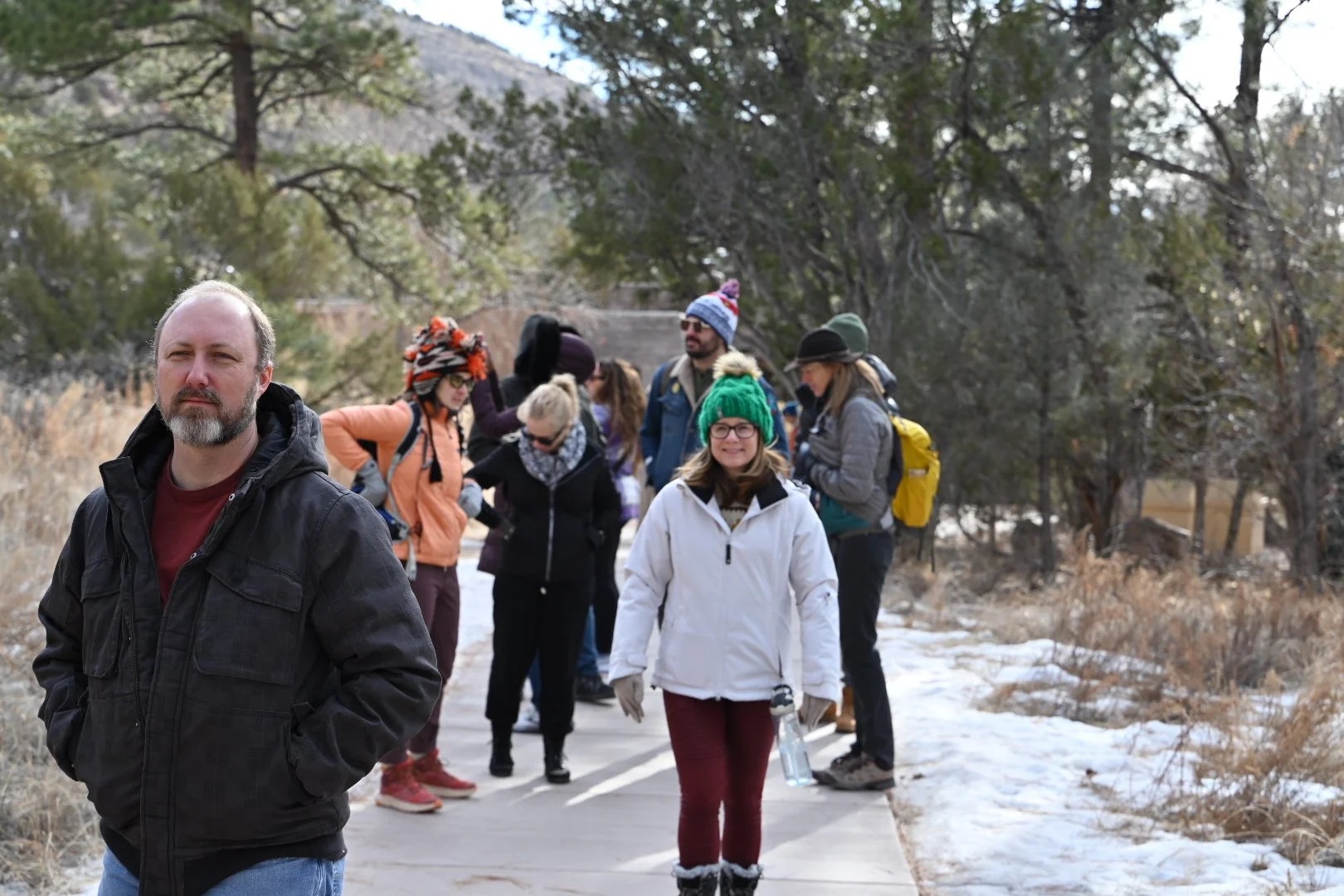 A group of people dressed for cold weather, walking on a snow-dusted path in a forested area.