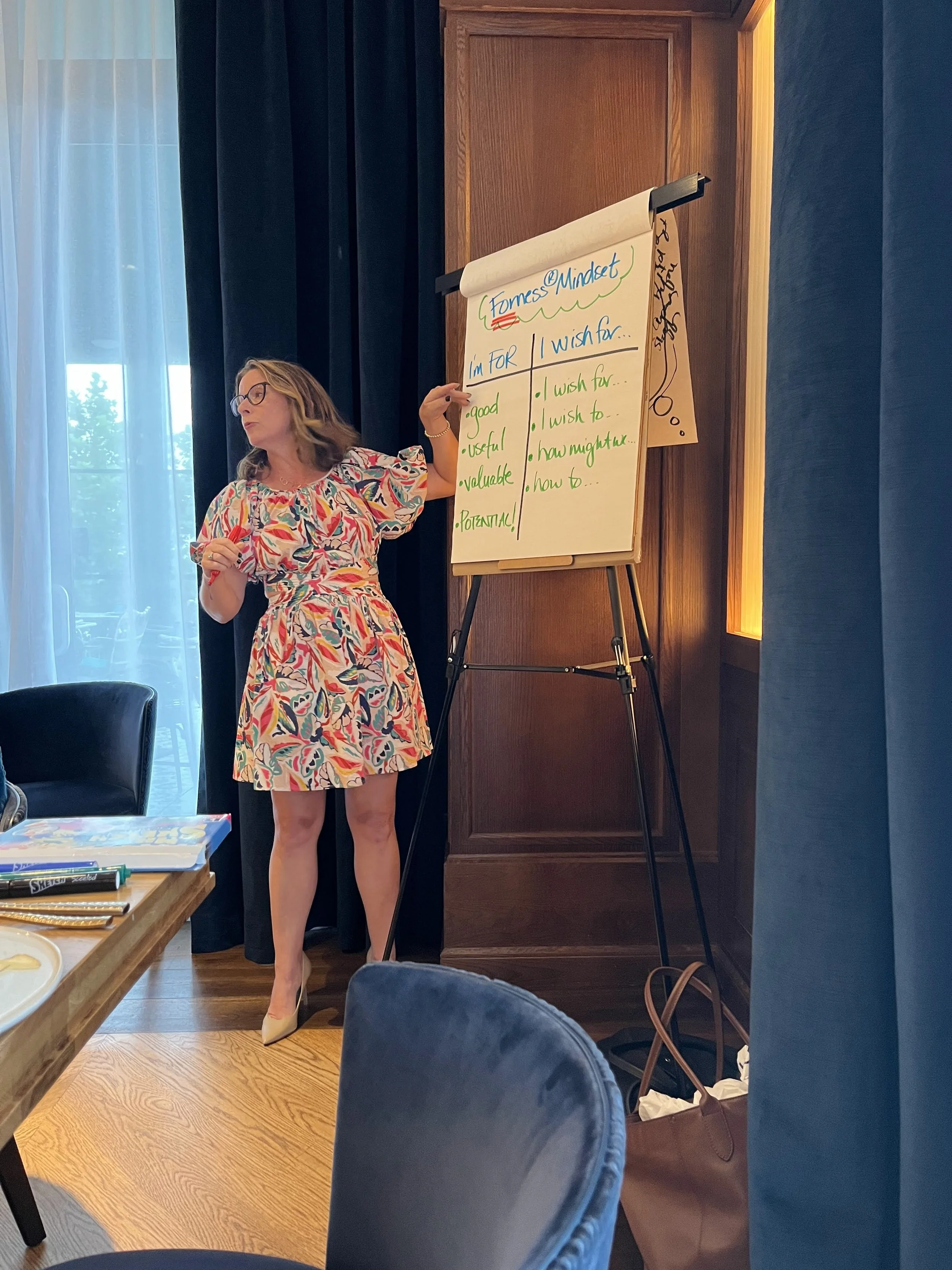 A woman with glasses wearing a colorful patterned dress, standing next to a flip chart, giving a presentation. The flip chart has handwritten notes about mindset and wishes, with topics like usefulness, value, and positivity.