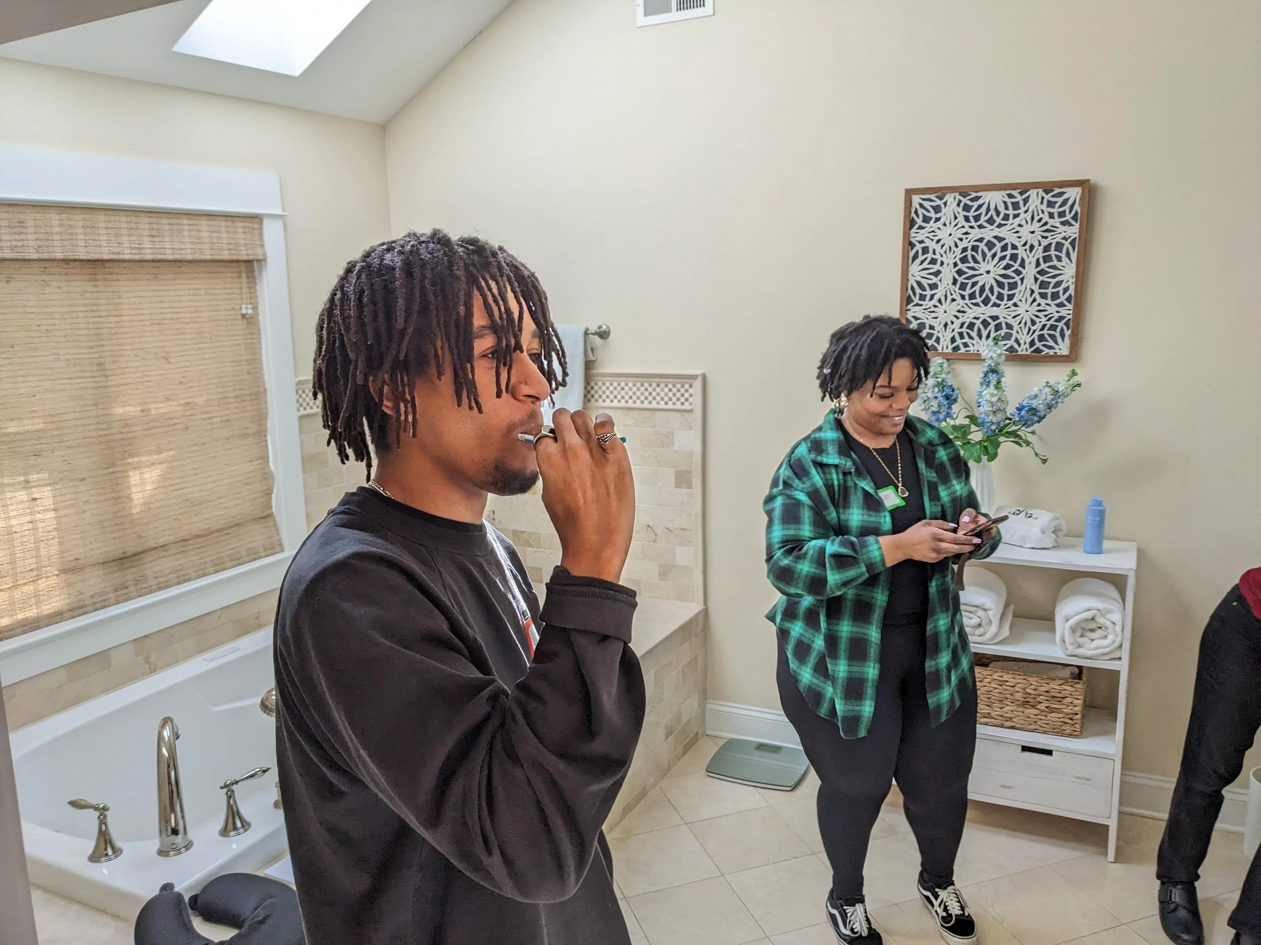 Consumer immersion with ITG. Two people in a bathroom; one is using a toothbrush, and the other is looking at a phone. The bathroom has a bathtub, a shelf with towels, a decorative wall piece, and a window with blinds.
