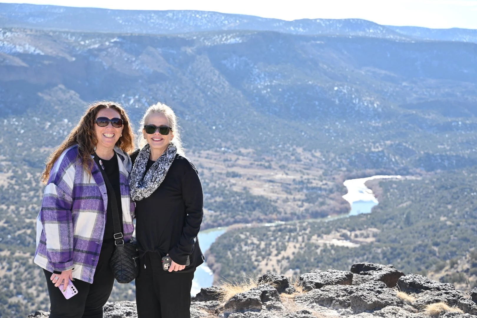 Two women smiling with sunglasses, standing on rocky terrain with a scenic view of mountains and a river in the background on a sunny day.