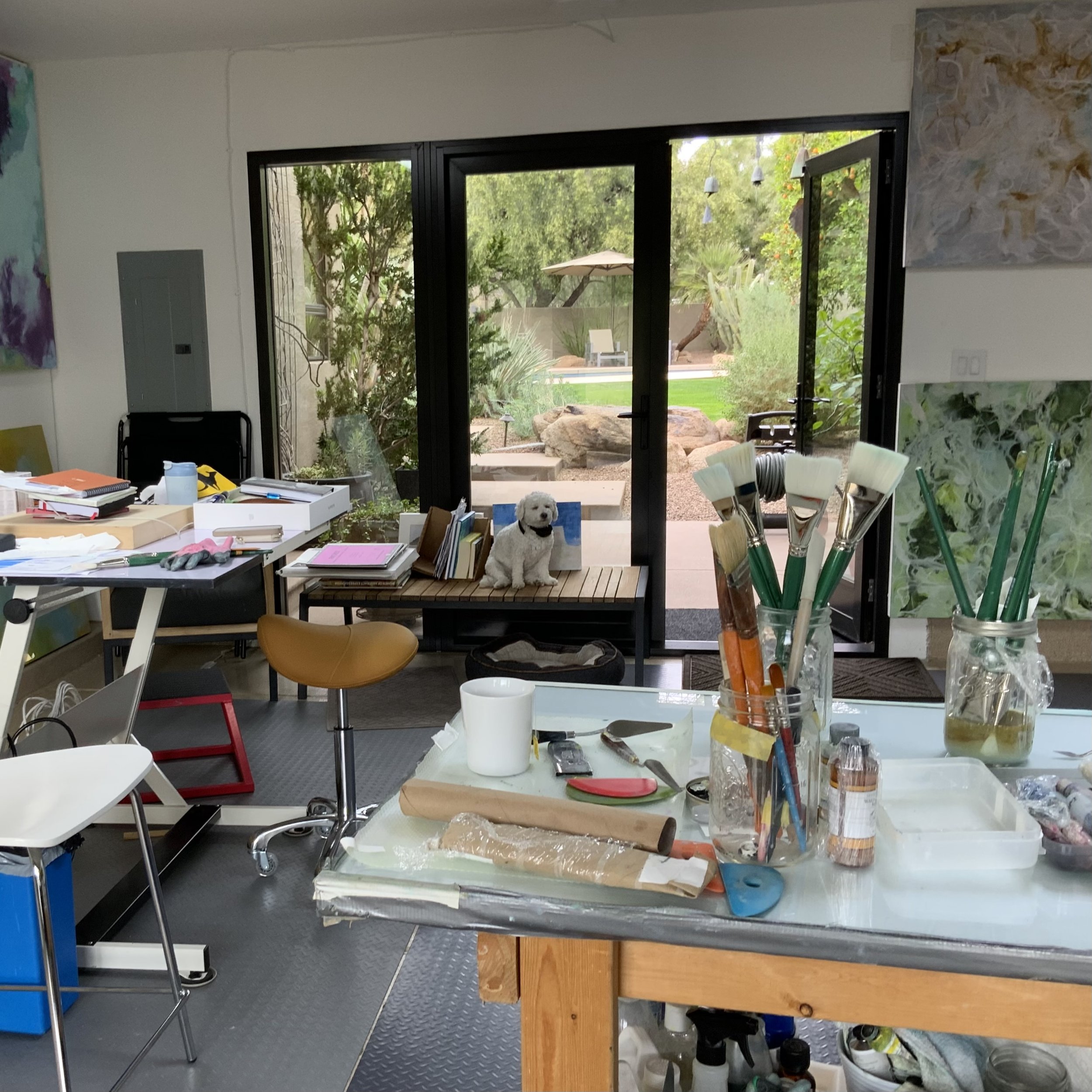 An indoor workspace with a table cluttered with papers, supplies, and a white dog sitting on a shelf. There are chairs, paintbrushes in a jar, and a view of a lush outdoor garden through large sliding glass doors.