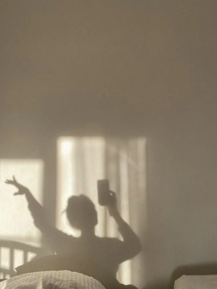 Person taking a mirror selfie in a dimly lit room, with curtains and a bed visible in the background.