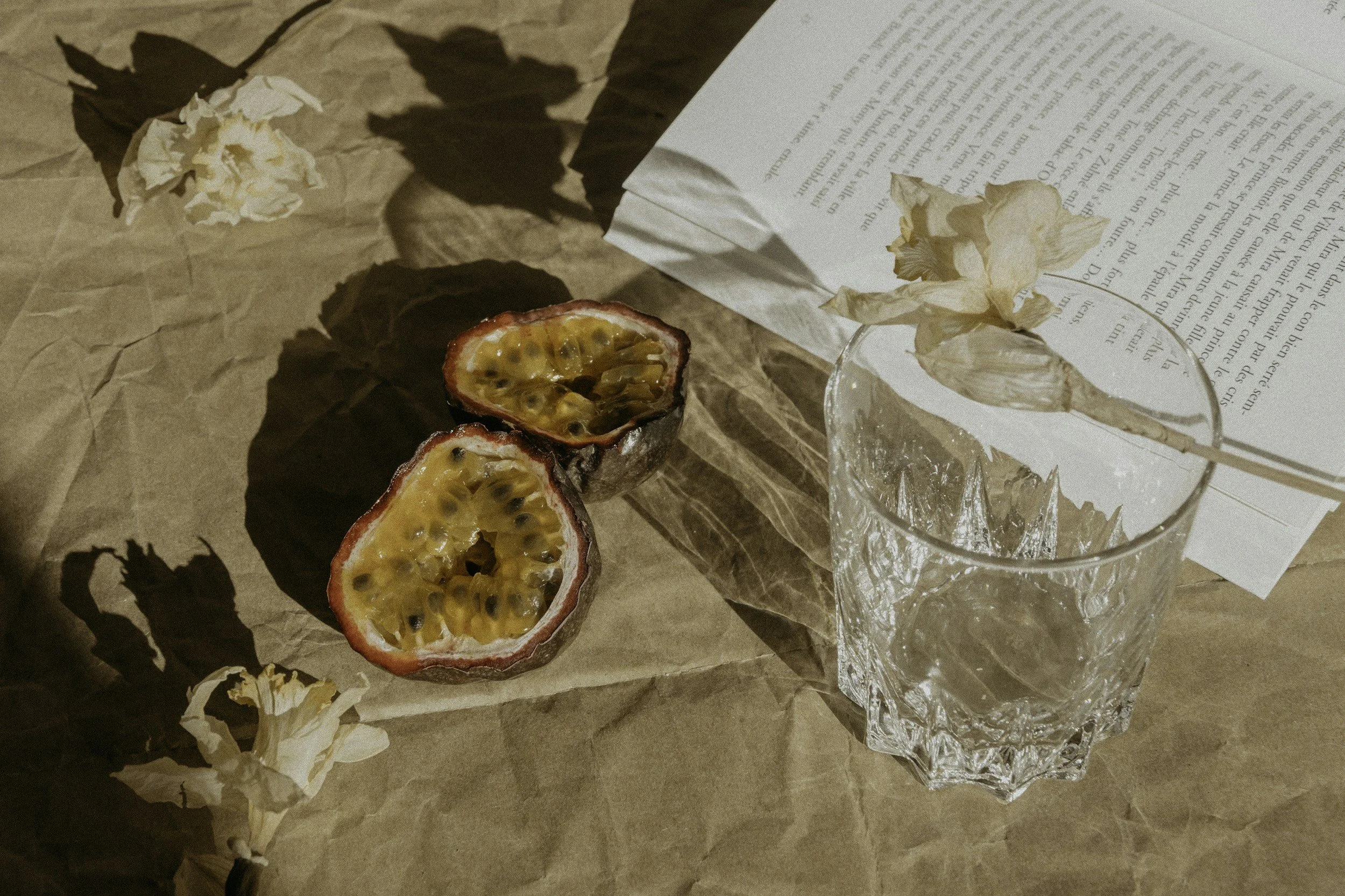 Halved passion fruit on crumpled brown paper with dried flowers, a glass with dried flowers inside, and a partially visible book with text