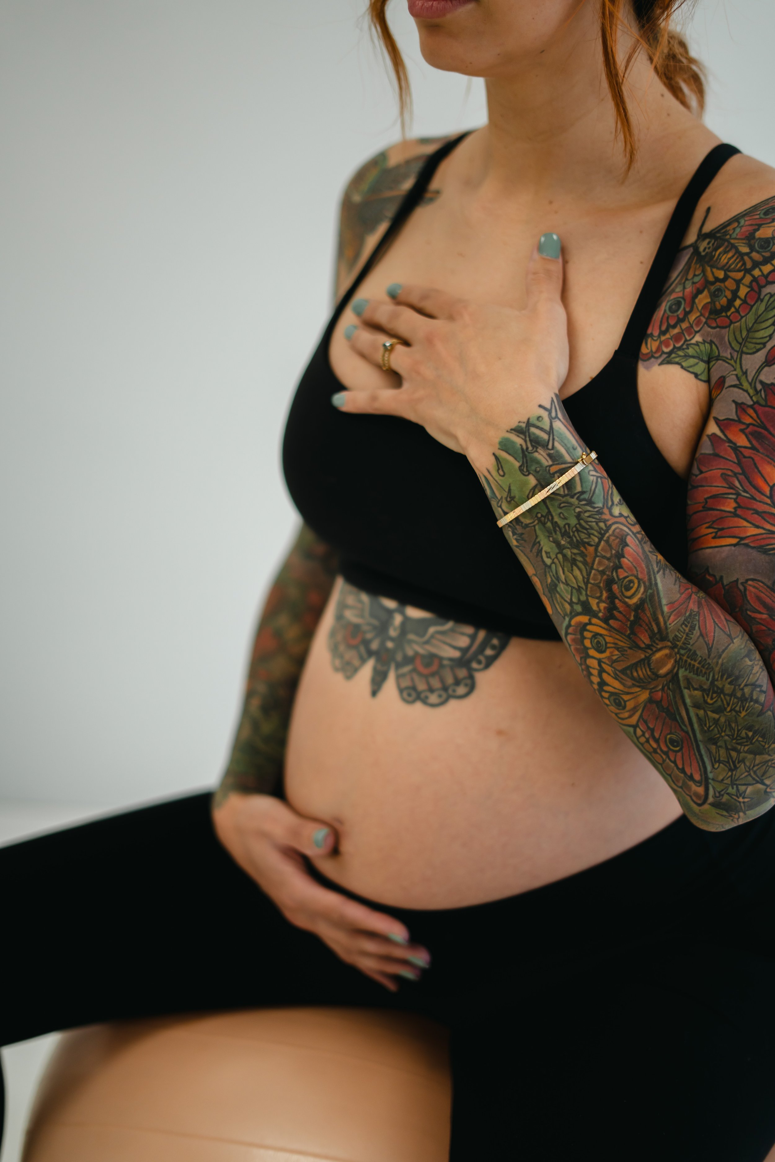 A pregnant woman with tattoos on her arms and torso, wearing a black tank top and black shorts, holding her pregnant belly with one hand, and touching her chest with the other.