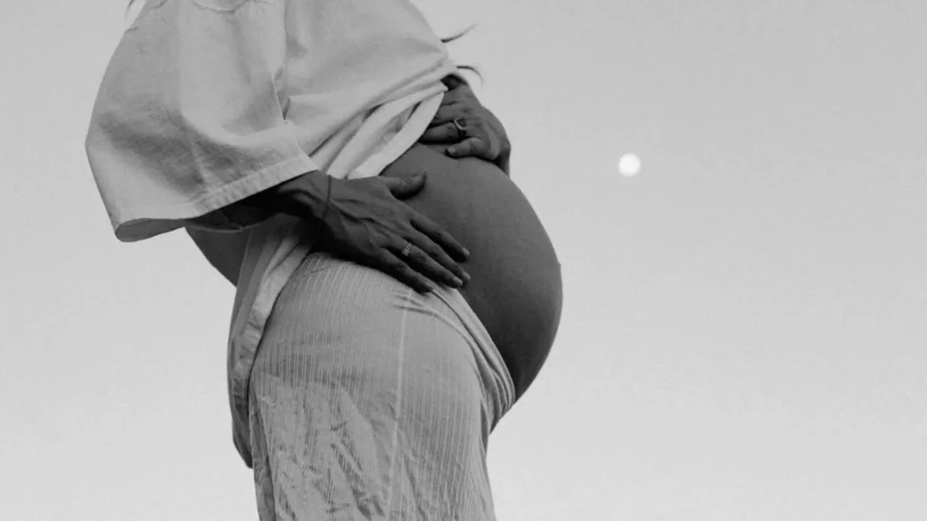 A pregnant person holding their belly outdoors during sunset or sunrise.
