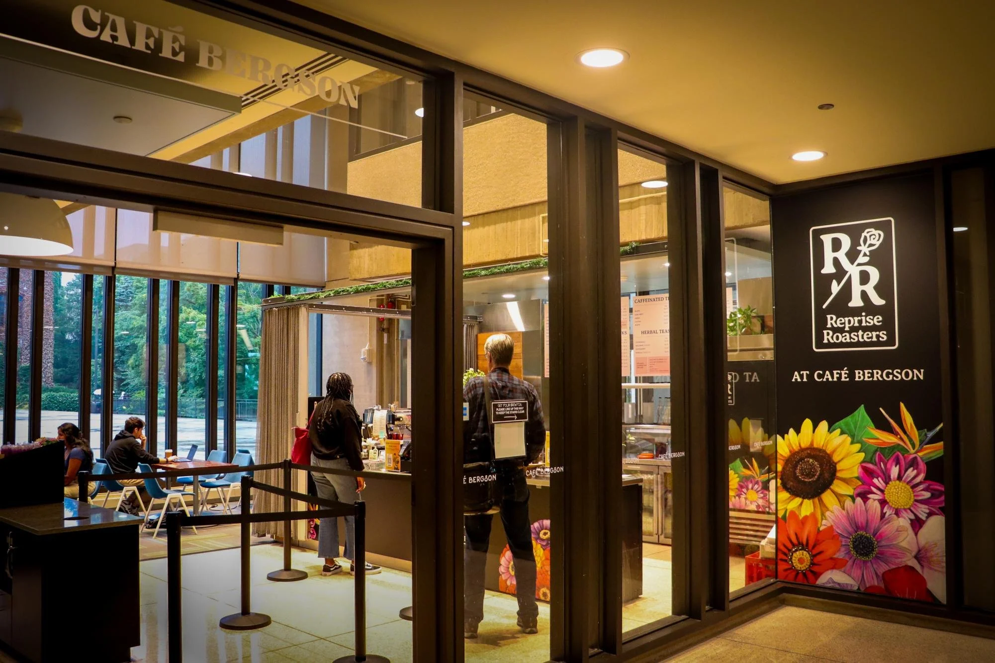 Interior of a cafe named Reprise Roasters with customers ordering at the counter, large windows with a view of greenery outside, colorful flower images on the wall, and warm lighting.