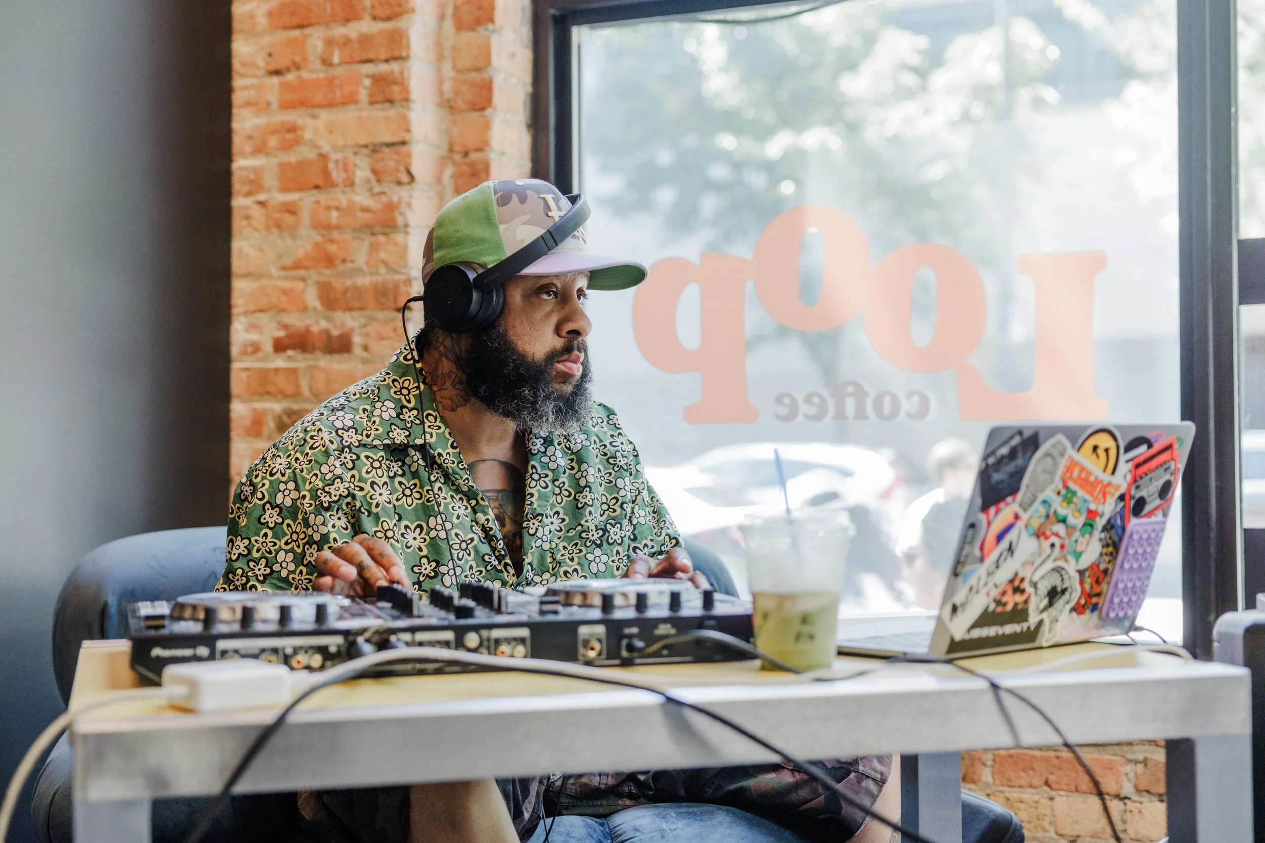 A man with a beard, tattoos, and wearing a colorful floral Hawaiian shirt with a camouflage cap, headphones, and tattoos on his neck, sitting at a table with DJ equipment and a laptop decorated with stickers, working or DJing in a cafe with a brick wall and large window in the background.