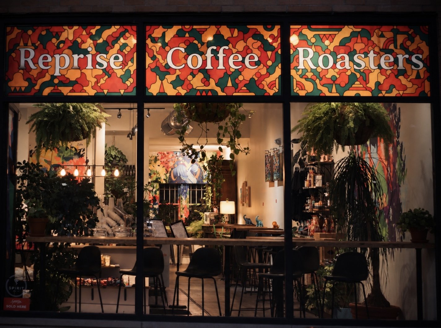 A storefront window of Reprise Coffee Roasters displaying colorful geometric patterns and inside, lush plants, colorful artwork, tables, chairs, and warm lighting.