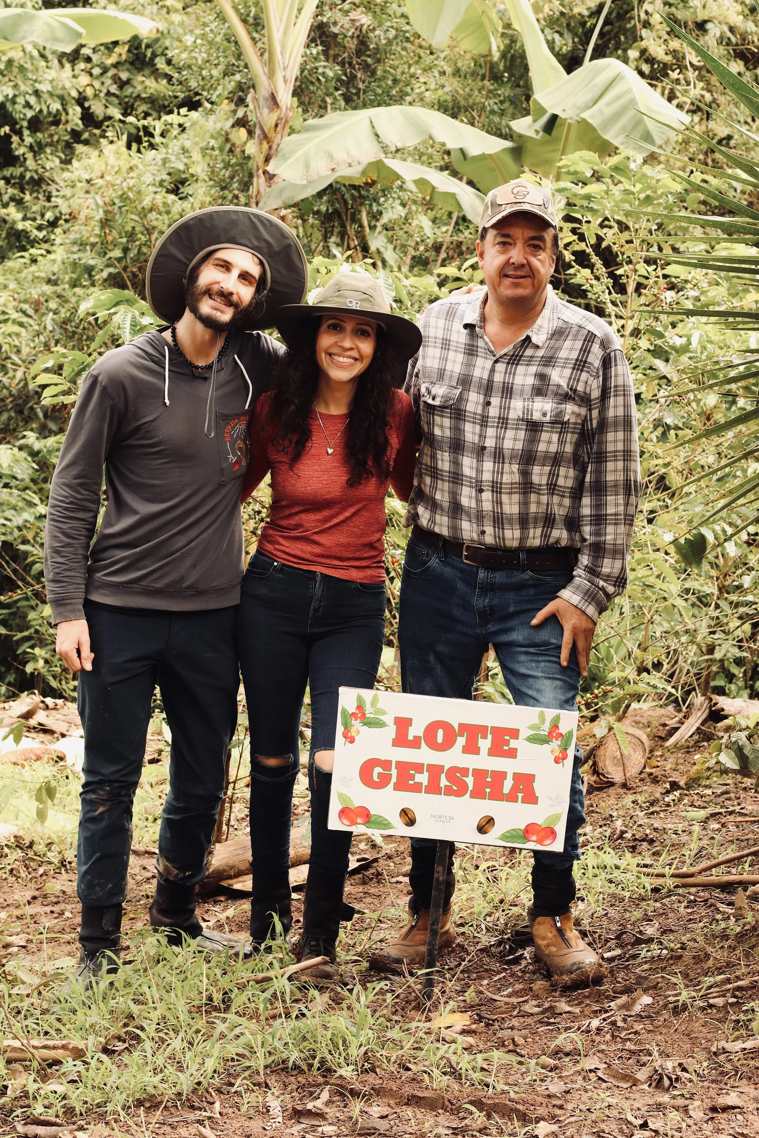 Three people standing outdoors in a jungle-like environment, holding a sign that reads "Lote Geisha," with a woman in the middle smiling and wearing a wide-brimmed hat. The man on the right is also smiling, wearing a plaid shirt and a cap. The man on the left has a beard, is smiling, and wearing a hoodie and a large hat. They are surrounded by lush green plants and large banana leaves.