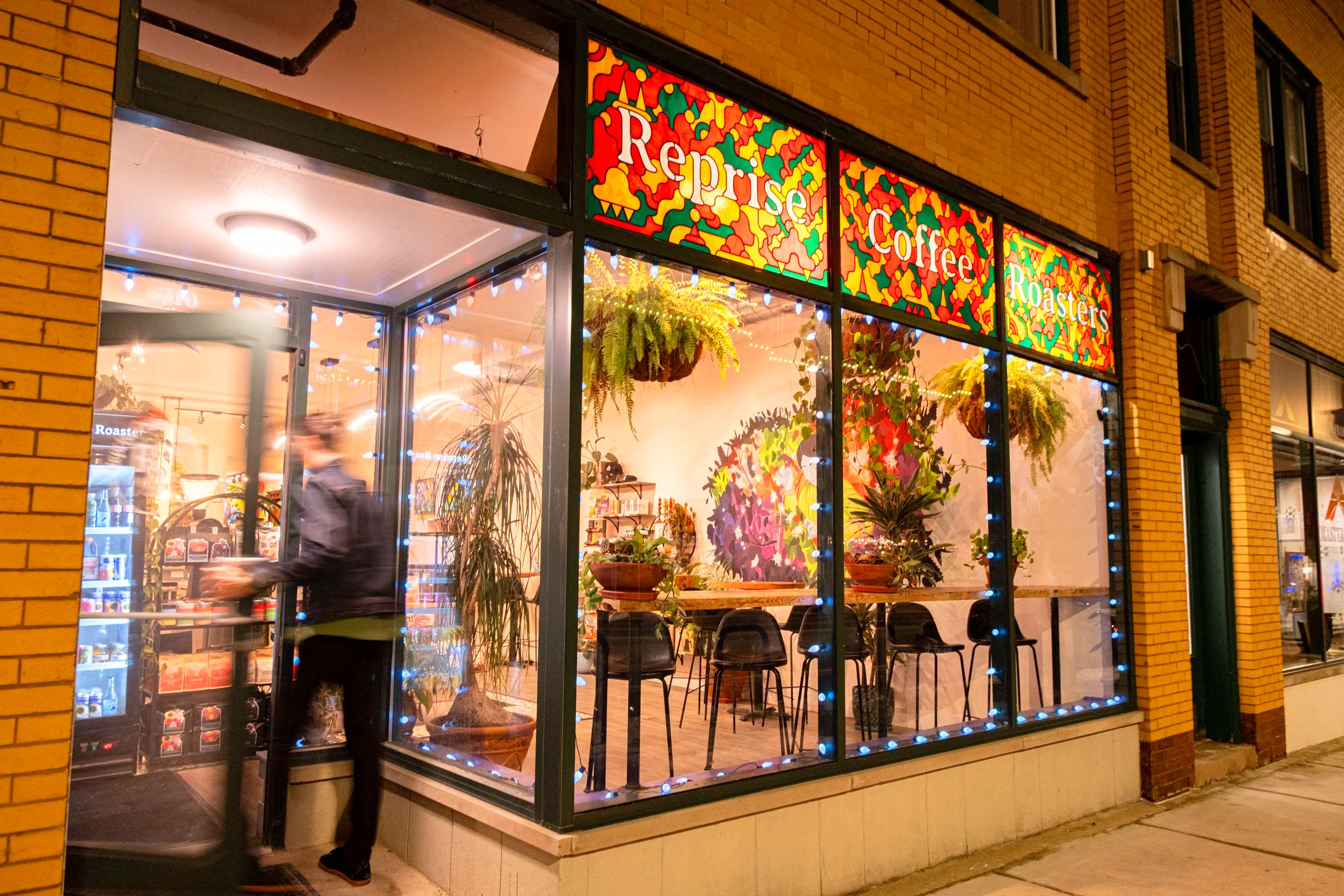 View of a storefront with large window display, colorful stained glass sign reading 'Reprise Coffee Roasters,' decorated with festive lights, inside has plants and seating, reflecting city street at night.
