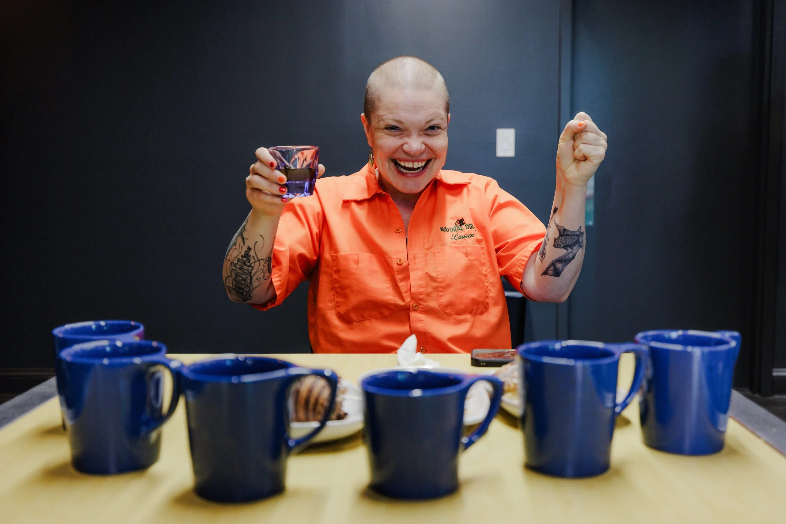 A person with a shaved head, tattoos, and earrings, wearing an orange shirt, smiling and holding a glass, surrounded by multiple ceramic mugs on a table.