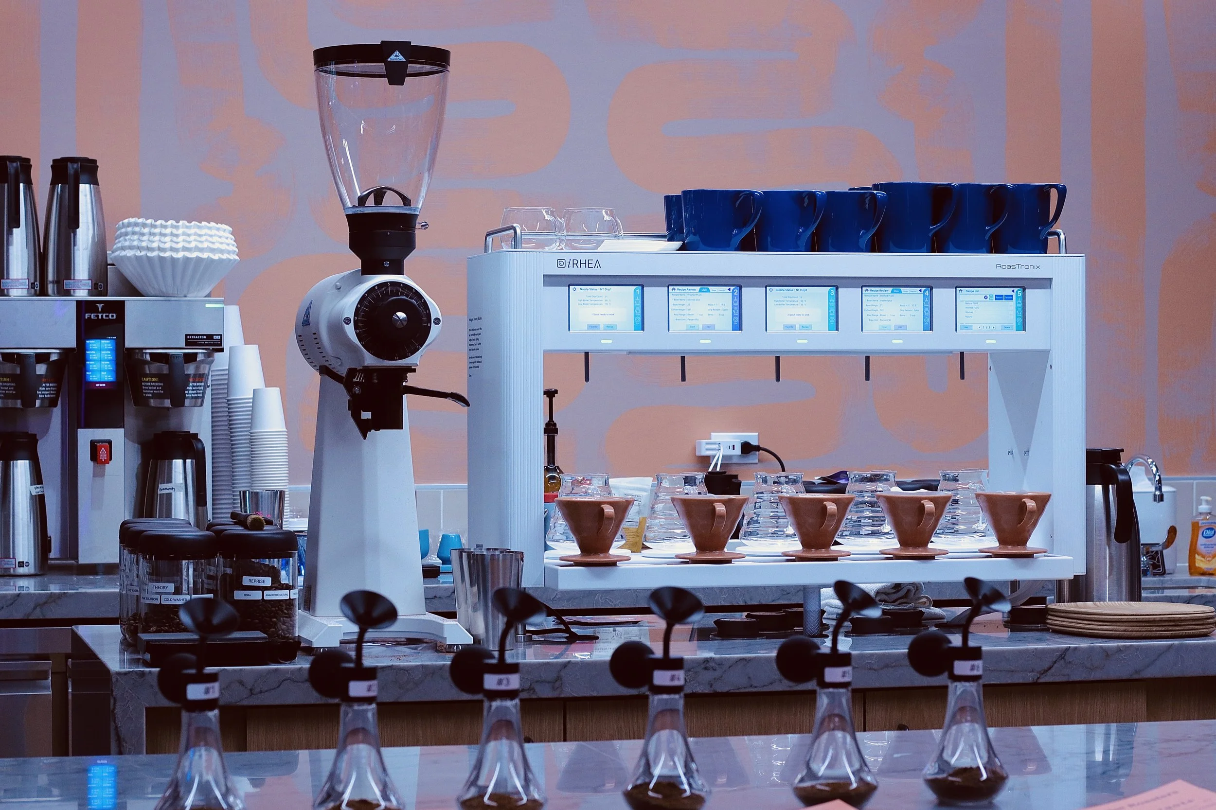 Coffee shop or cafe setup with brewing equipment, cups, and coffee filters on a counter, with a pink and lavender abstract patterned wall in the background.