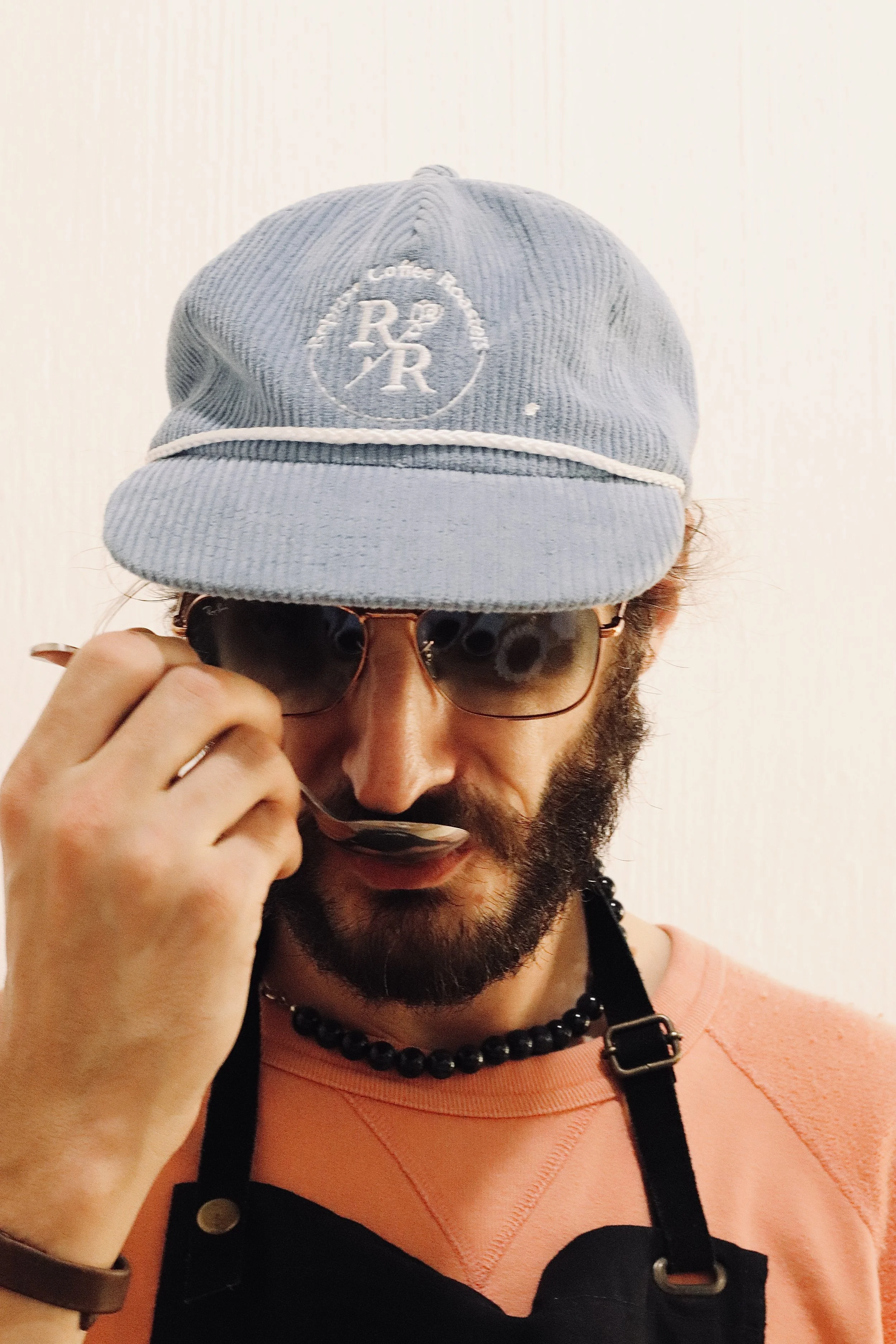 Person wearing a blue cap with logo and sunglasses, adjusting their sunglasses with their hand, wearing an orange shirt, black beaded necklace, and black apron.