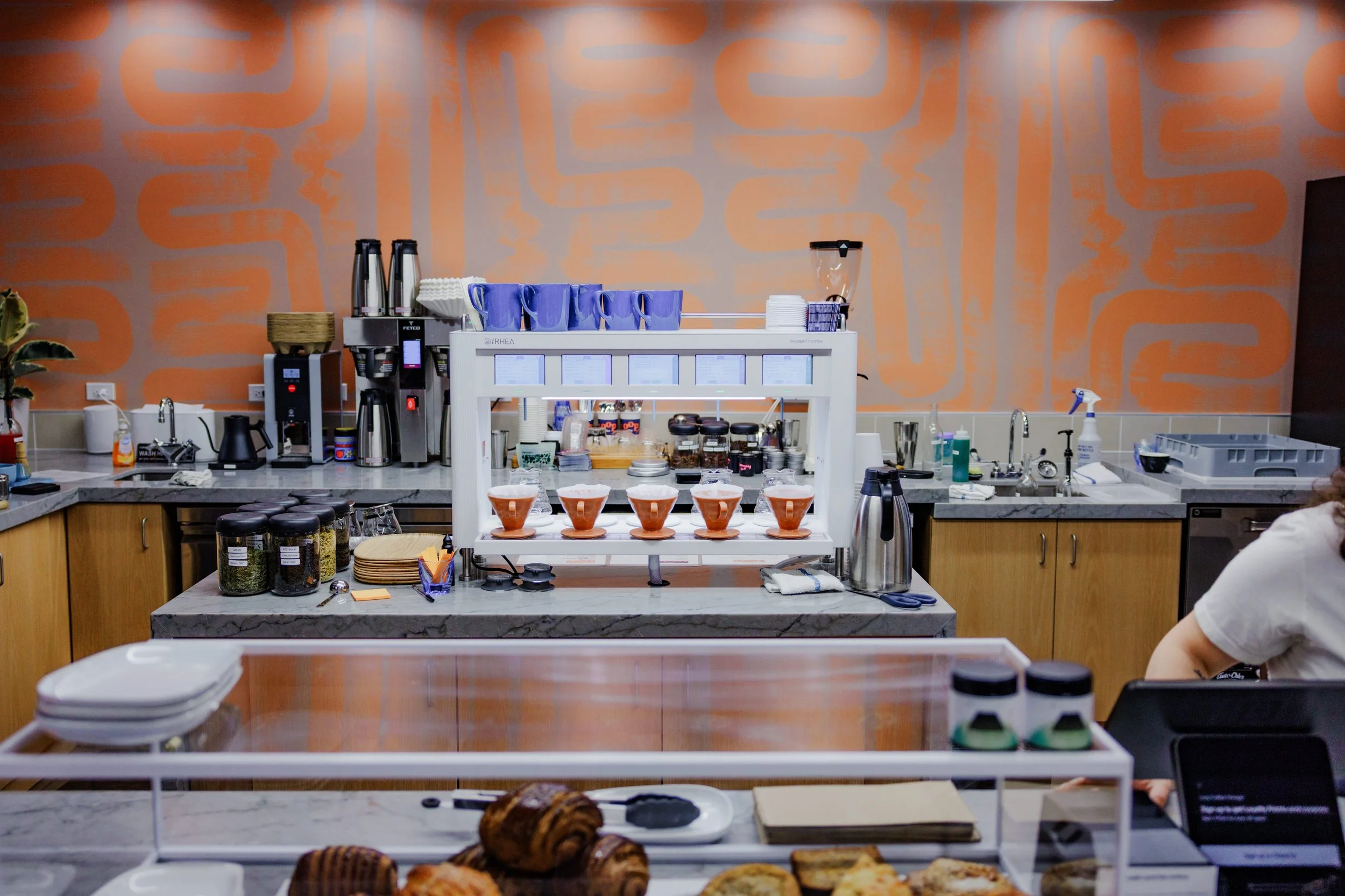 Coffee shop counter with cups, jars, kitchen equipment, pastries, and baked goods against an orange patterned wall.