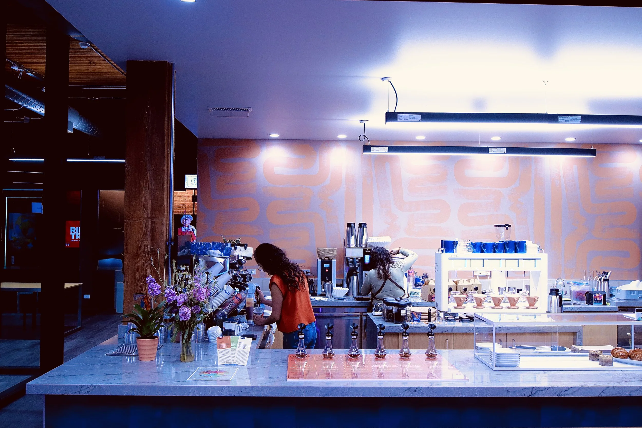 Interior of a modern coffee shop or café with people working behind the counter, floral arrangements, and coffee preparation equipment.