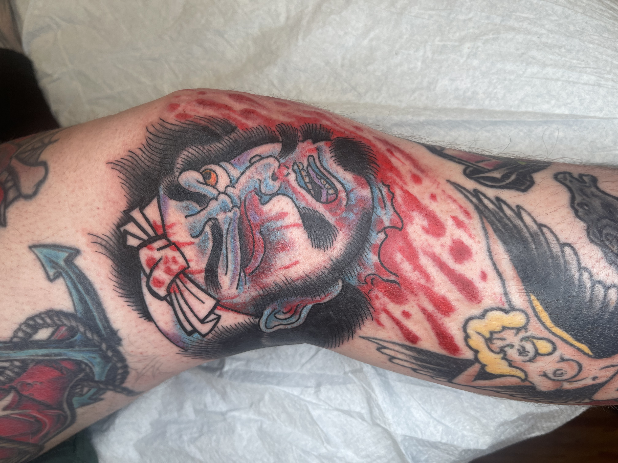 A tattoo on a person's arm featuring a colorful skull with black hair, surrounded by red ink splatters and other intricate designs.